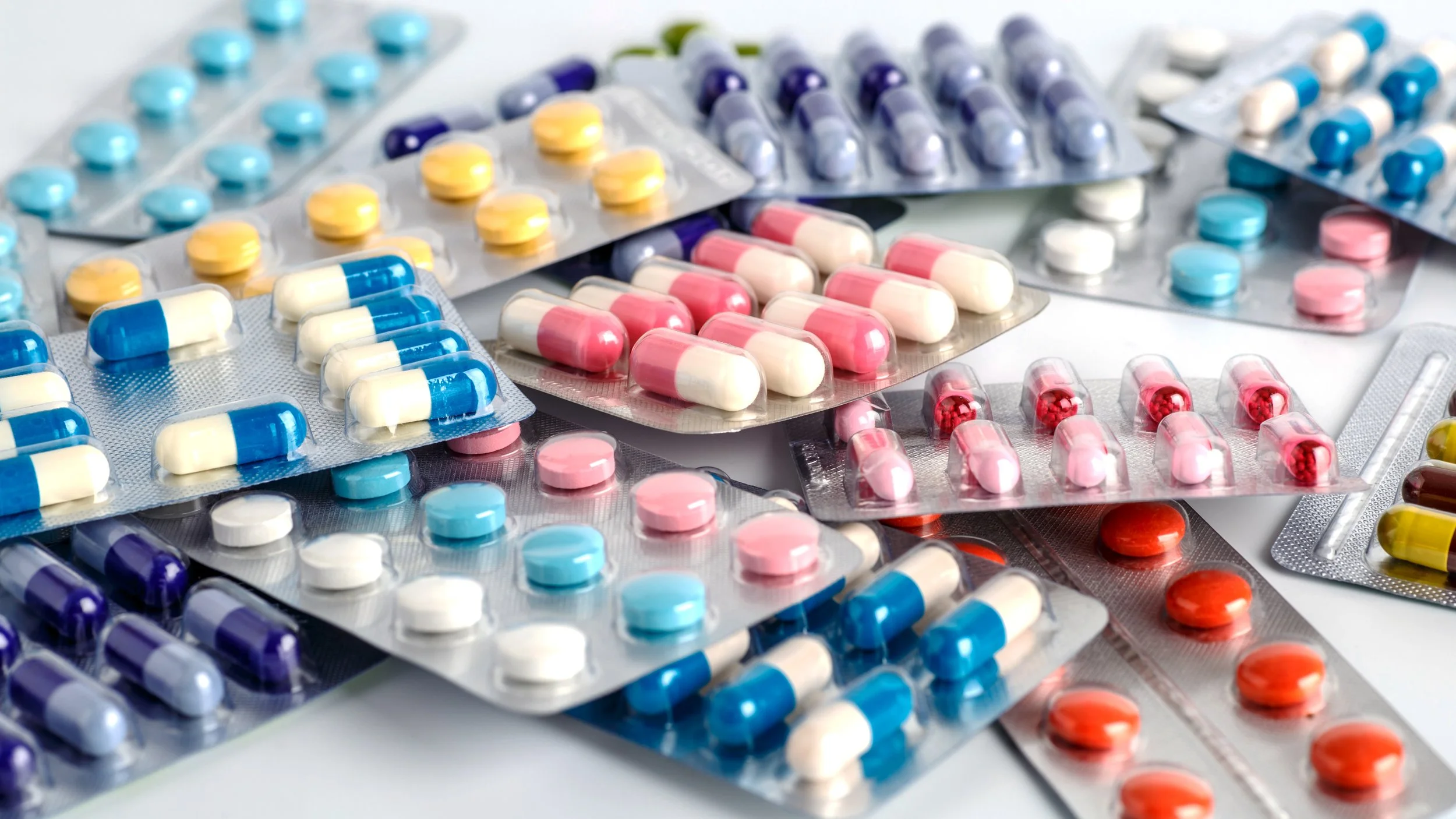 Multiple blister packs of pills and capsules in various colors and sizes scattered on a white surface.
