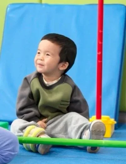 Child sitting on the floor indoors, smiling, with blue and green padded mats and play equipment around him.