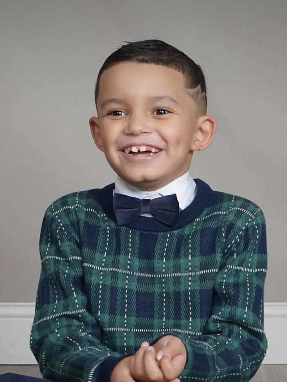 Young boy with a gap-tooth smile, wearing a green and navy plaid sweater, white shirt, and navy bow tie, posing against a neutral gray background.