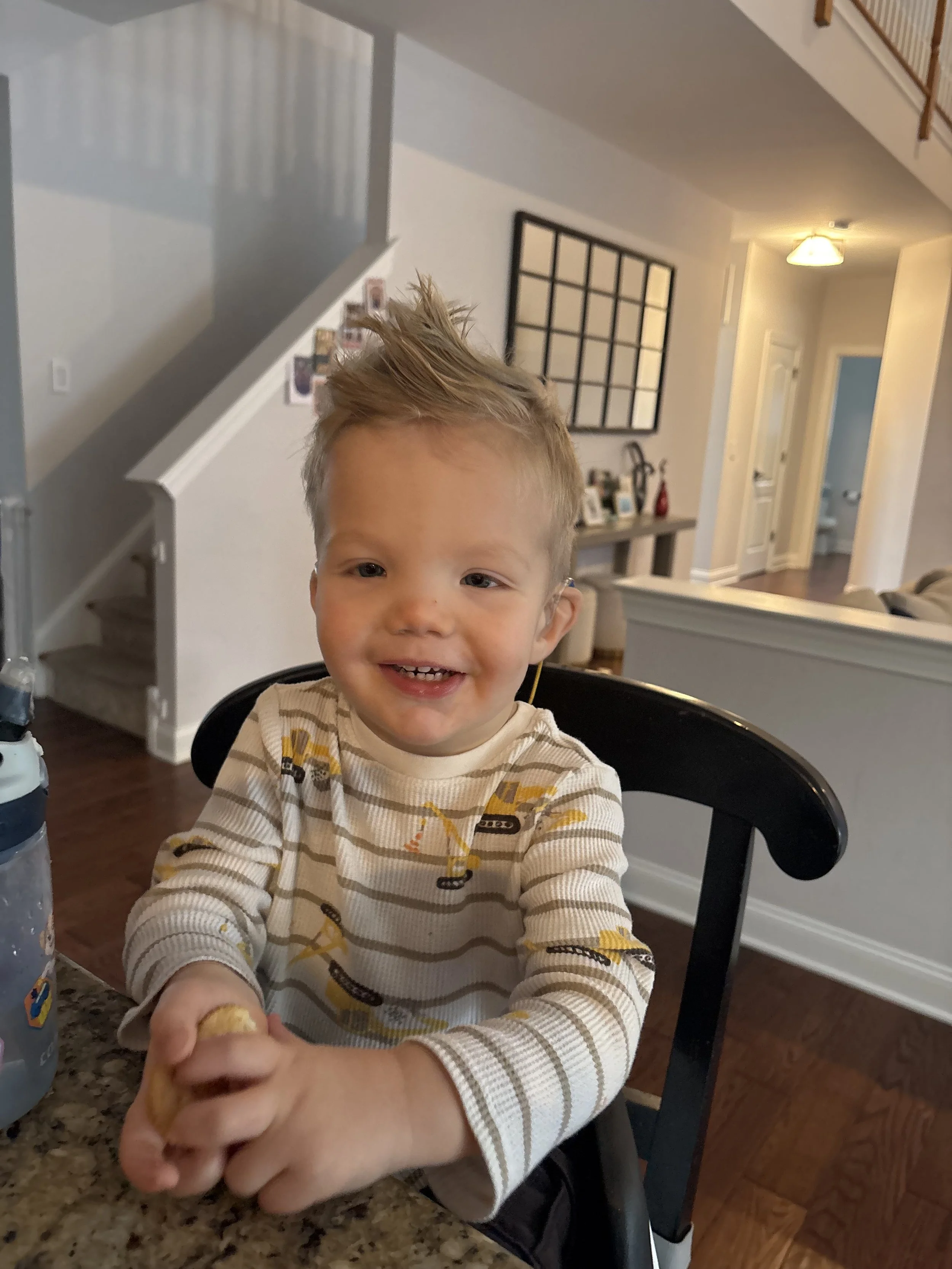 A young boy with spiky blonde hair and a striped cream and yellow shirt is sitting in a high chair at a kitchen counter, smiling.