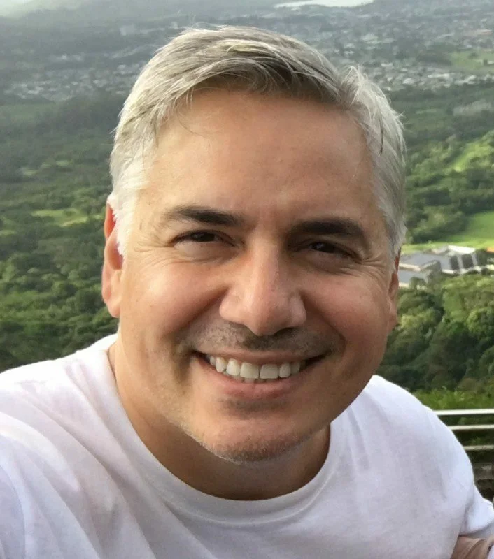 A smiling, handsome man with gray hair wearing a white t-shirt in a green, outdoor landscape.
