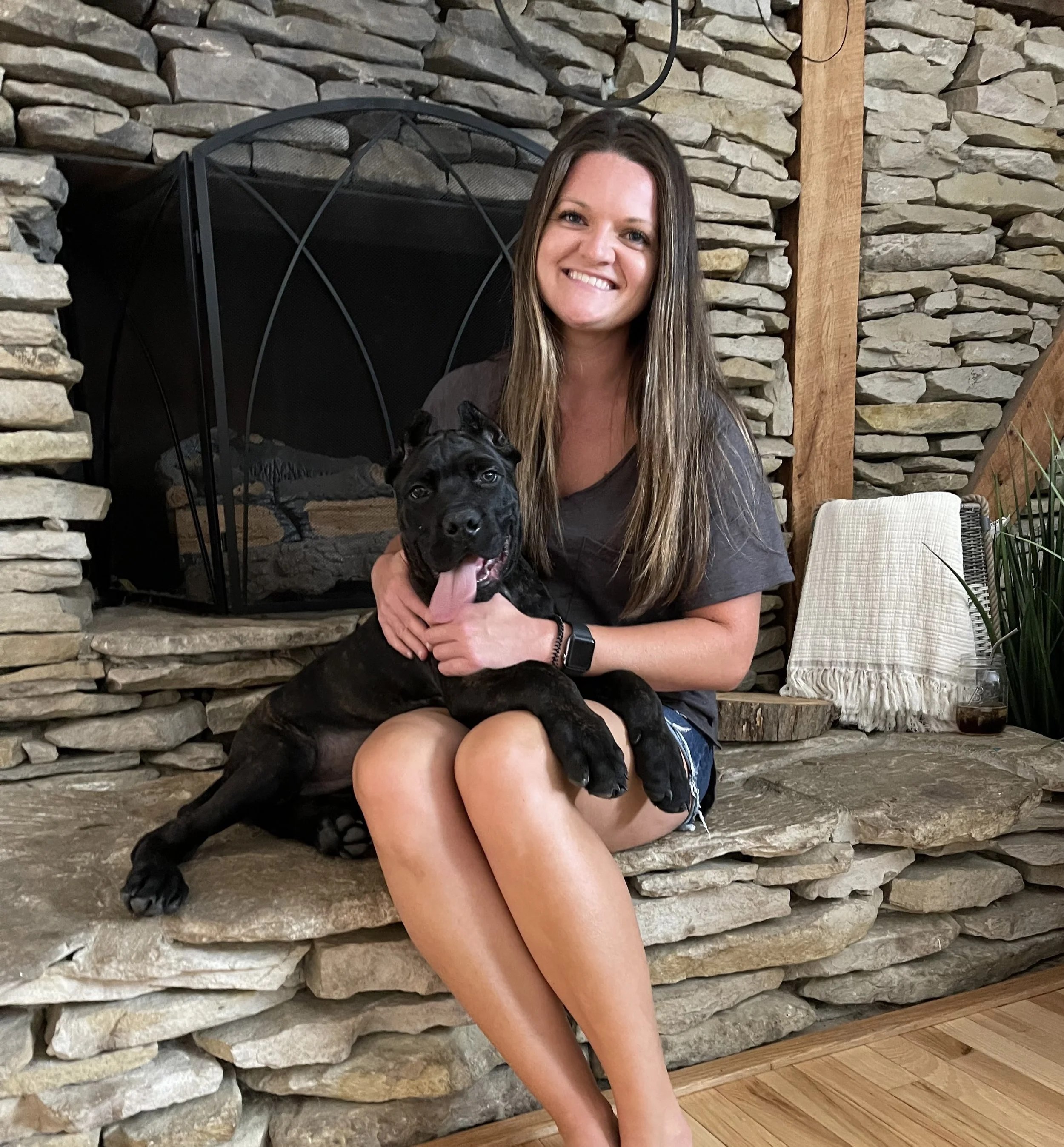 A woman with long brown hair and wearing a dark T-shirt sitting on a stone hearth, holding a young black puppy with a brindle coat and pink tongue, in front of a stone fireplace.