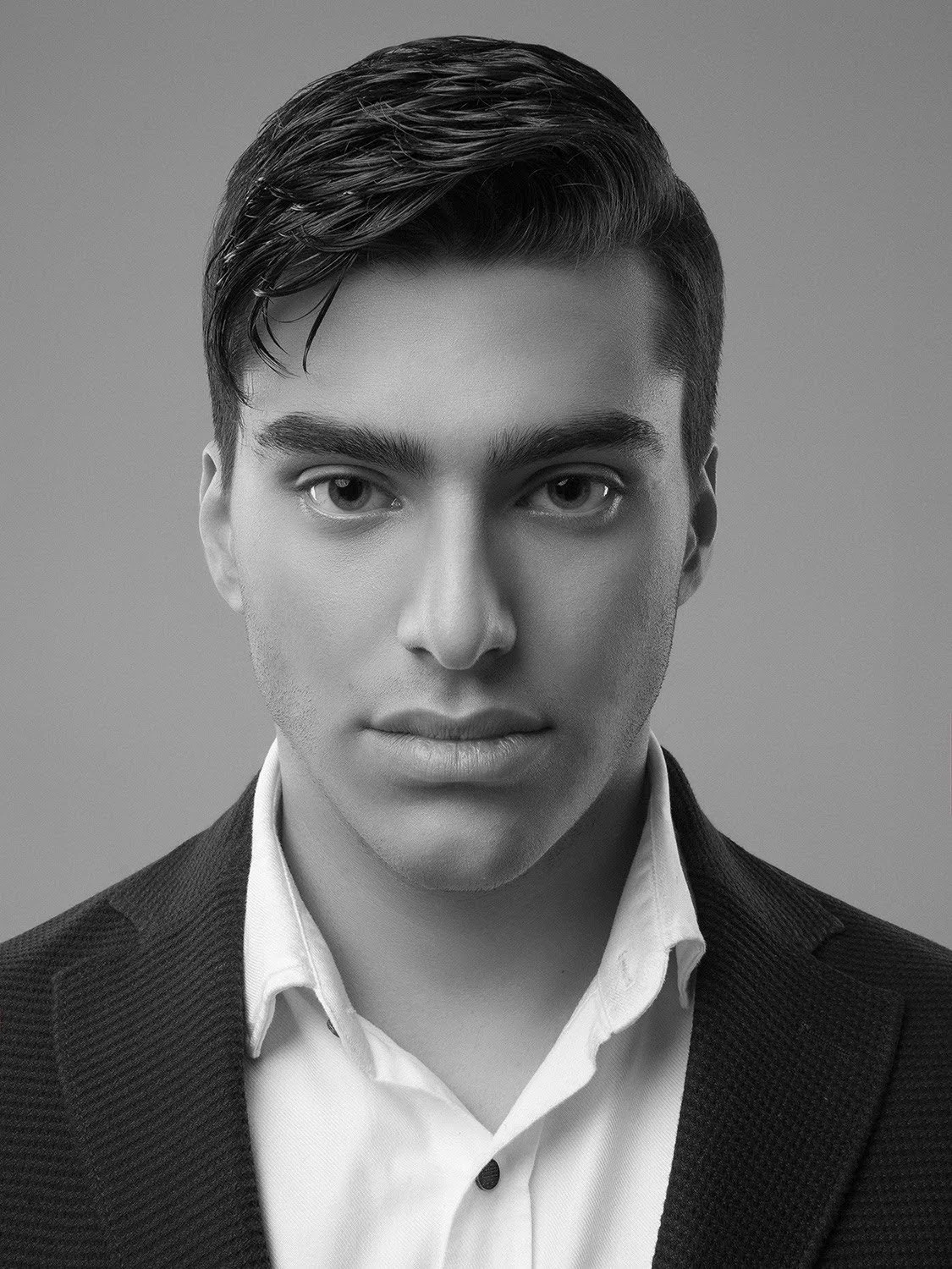 Black and white portrait of a young man with slicked-back dark hair, wearing a white shirt and a dark blazer, looking directly at the camera.