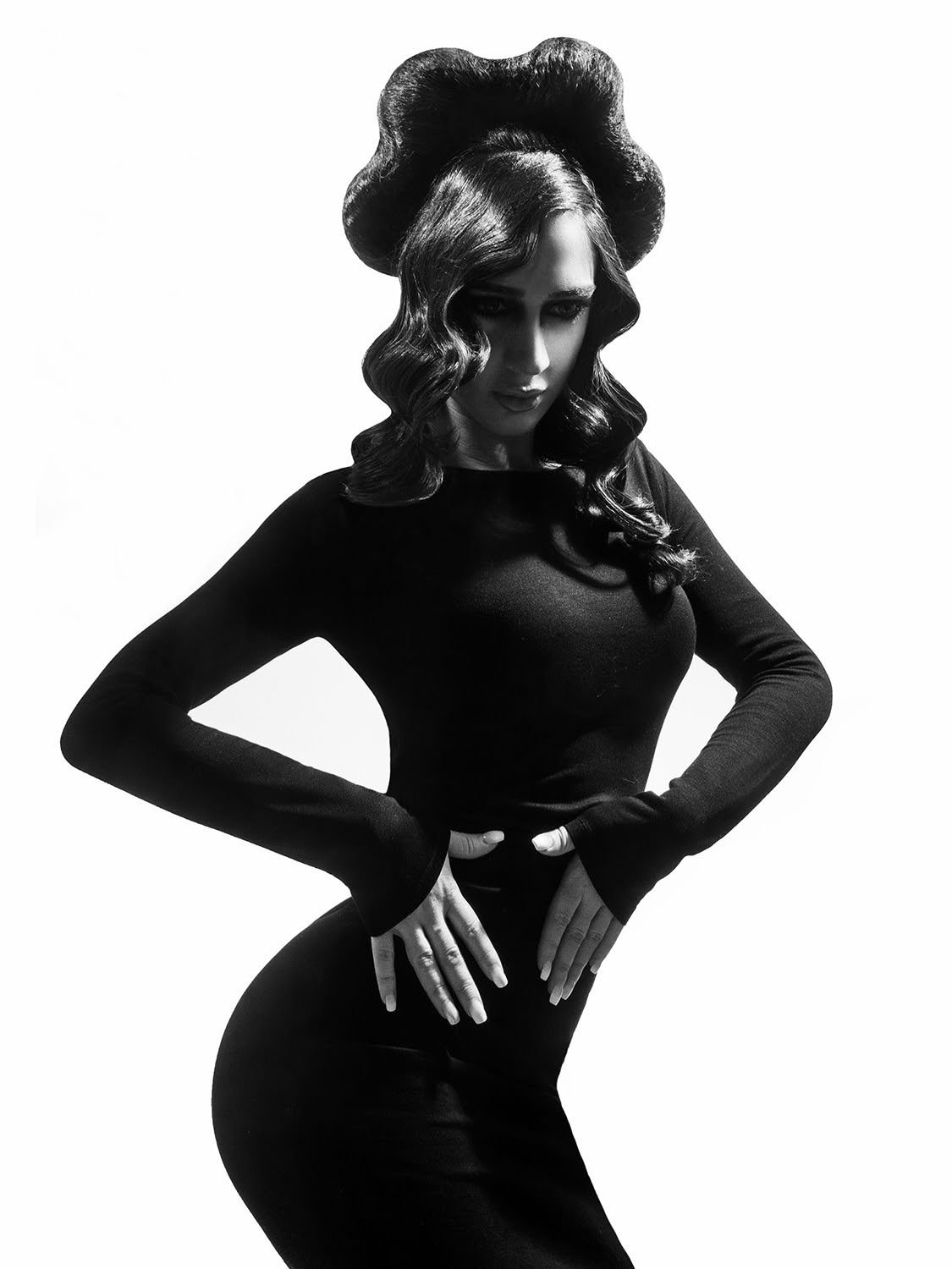 Black and white photo of a woman with styled hair, wearing a form-fitting long-sleeve top, posing with her hands on her hips against a plain background.
