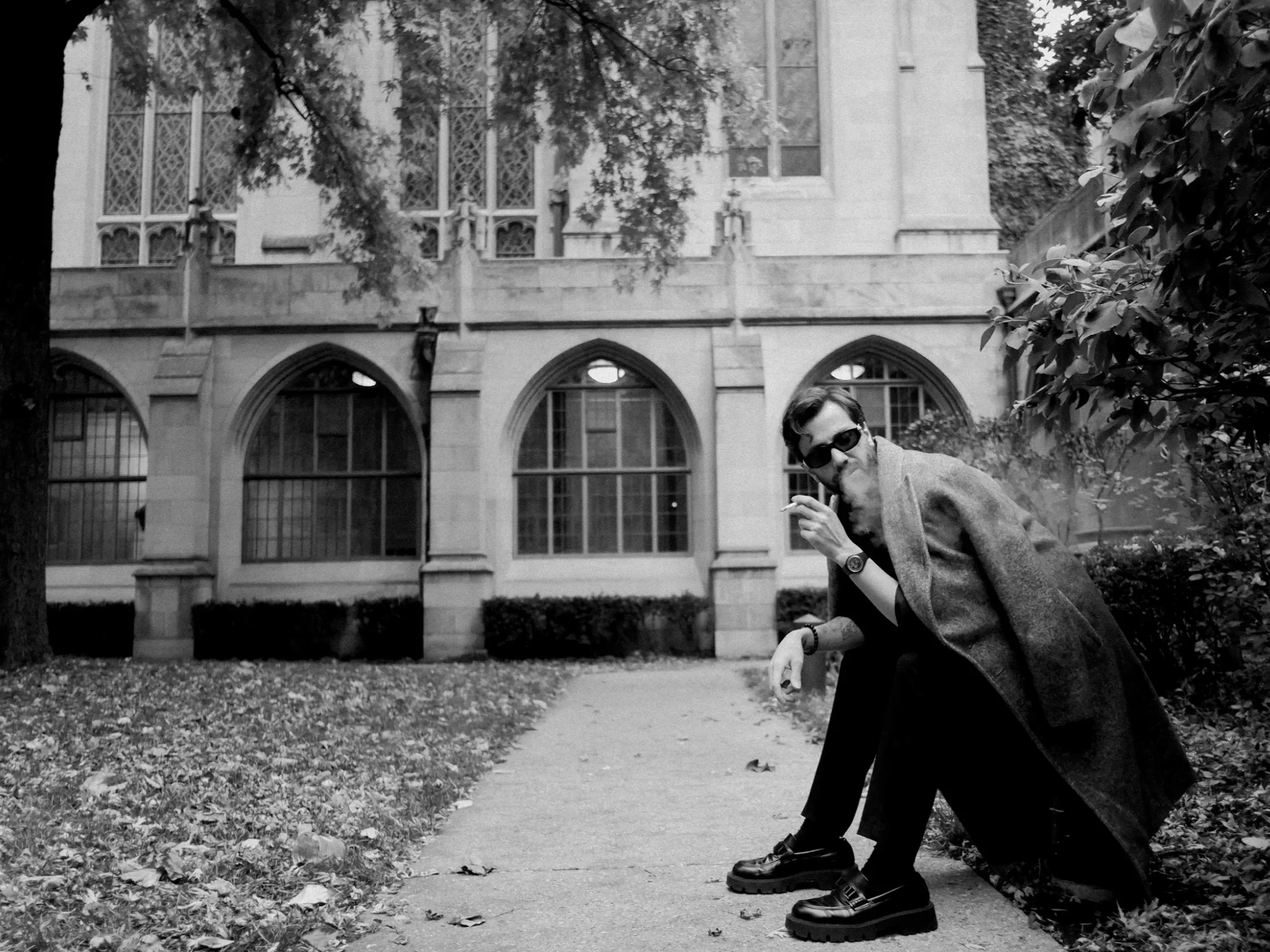 A man with sunglasses, smoking a cigarette, sitting on a park bench in front of a large stone building with arched windows, wearing a long coat and dark shoes, surrounded by trees and fallen leaves.
