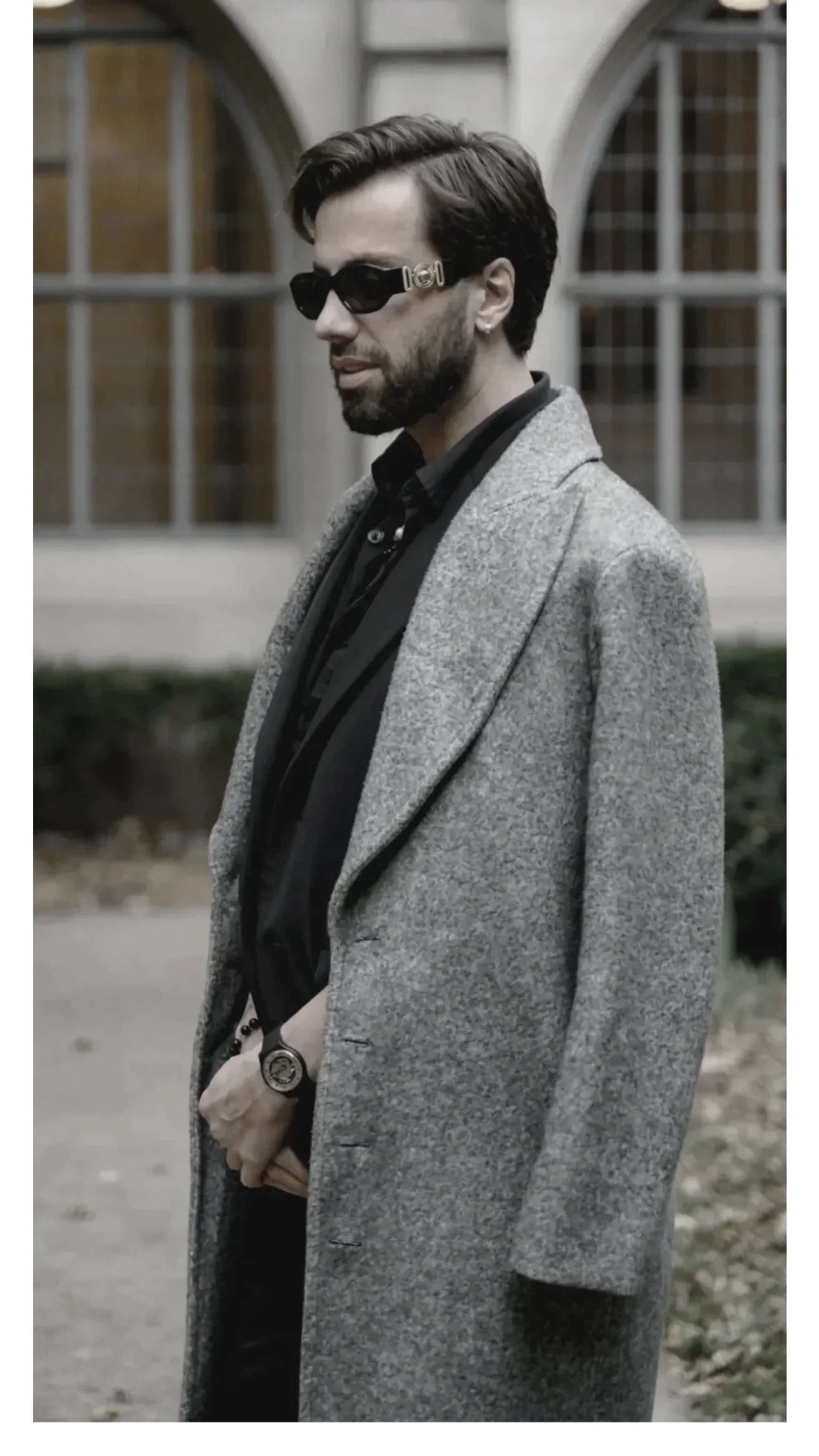 Portrait of a luxury hairstylist in a tailored gray coat and black shirt, standing in an urban setting, expressing confidence, precision, and modern style.