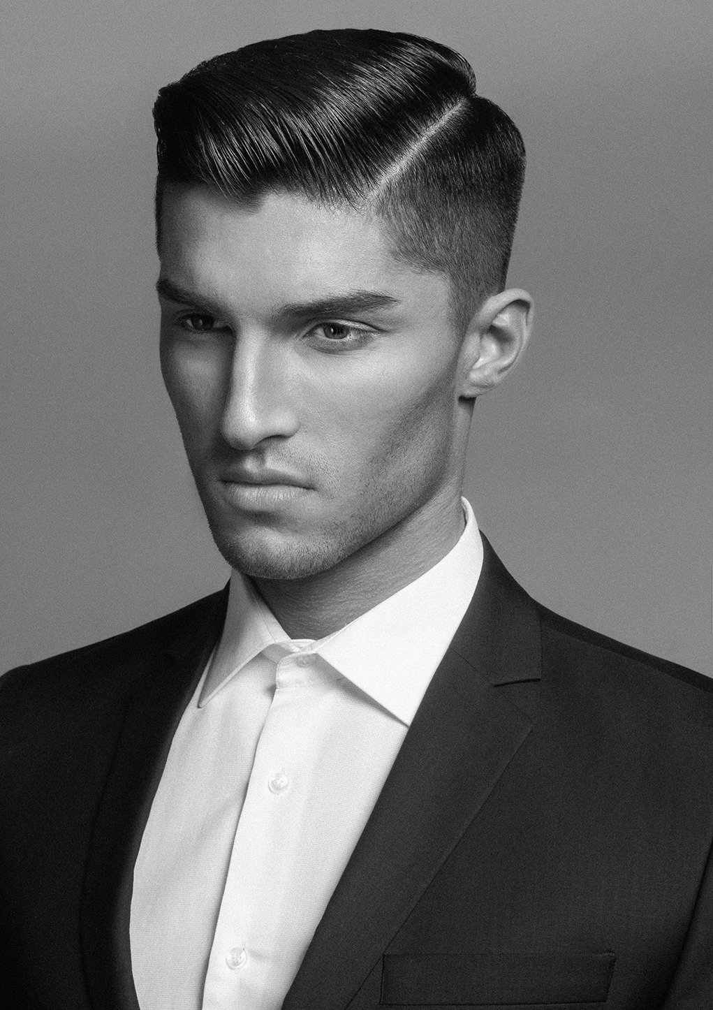 Black and white portrait of a young man dressed in a suit and a white shirt, with slicked-back hair, looking down with a serious expression.