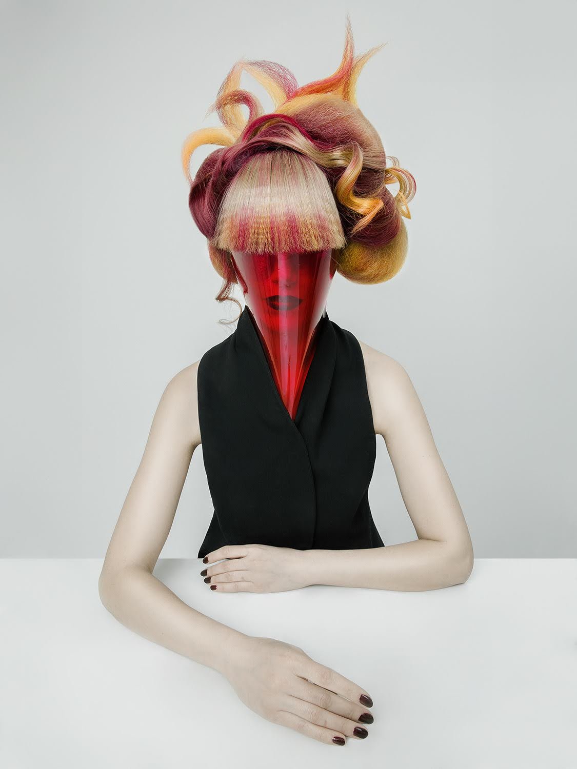A woman with a surreal hairstyle wears a black sleeveless top and a red, transparent mask covering her face, sitting at a white table against a plain background.
