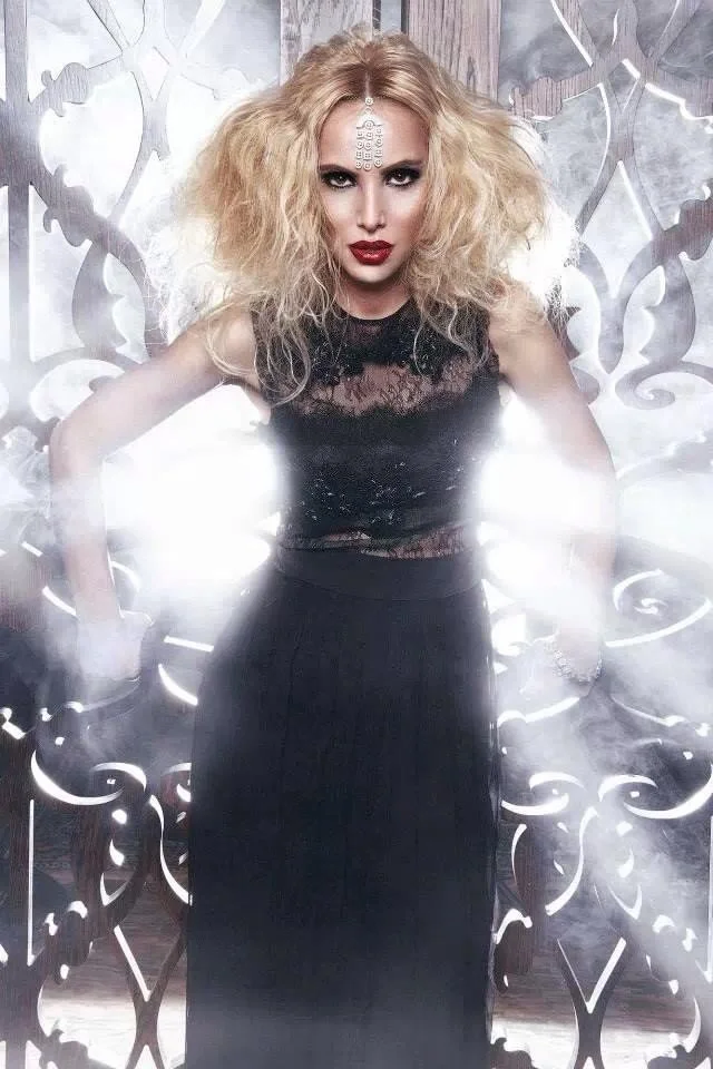 A woman with frizzy blonde hair, bold makeup, and red lipstick posing in front of an ornate metallic backdrop, wearing a black lace top and long skirt.