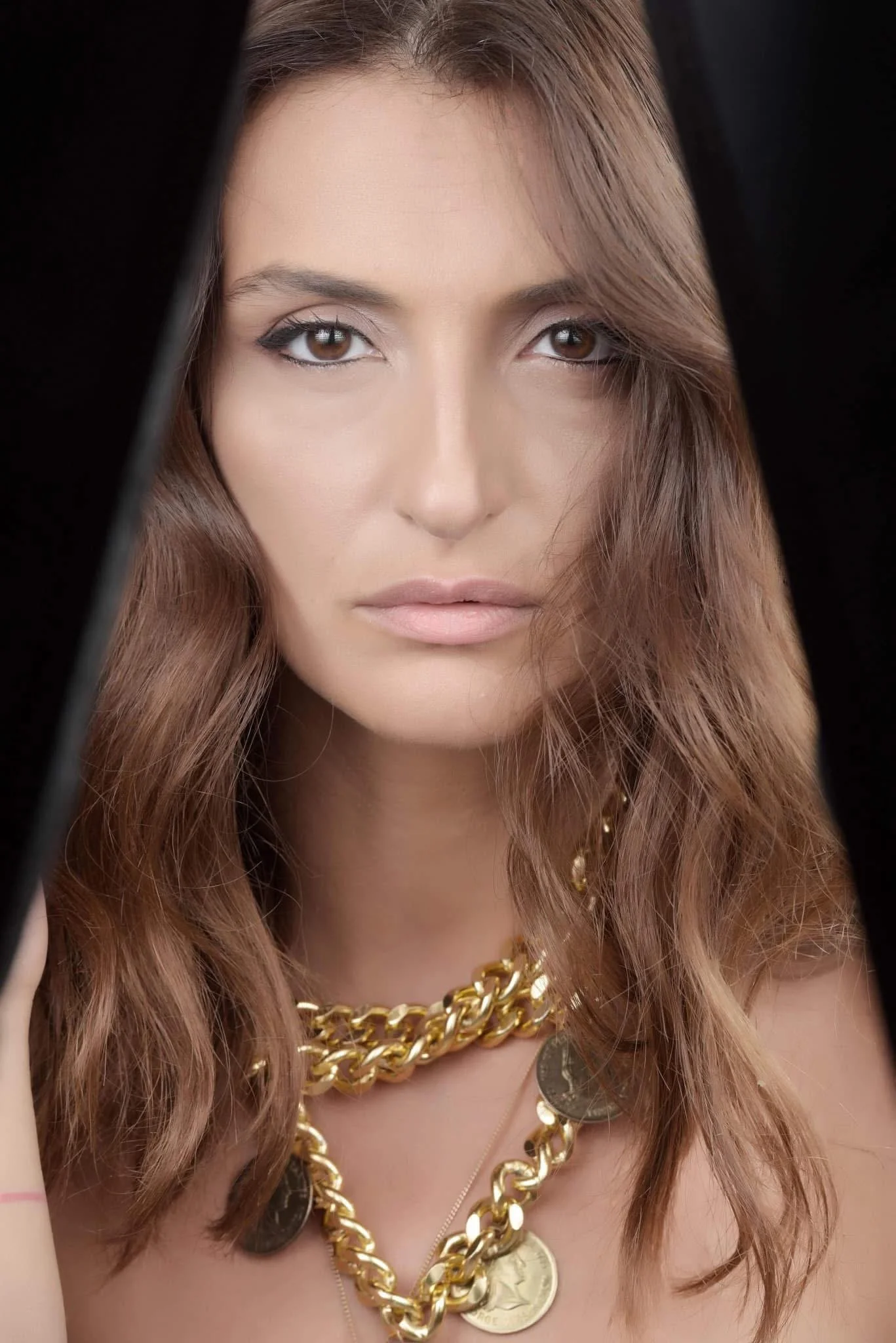 Close-up portrait of a woman with brown hair and light makeup, wearing multiple gold chains with coins, looking directly at the camera