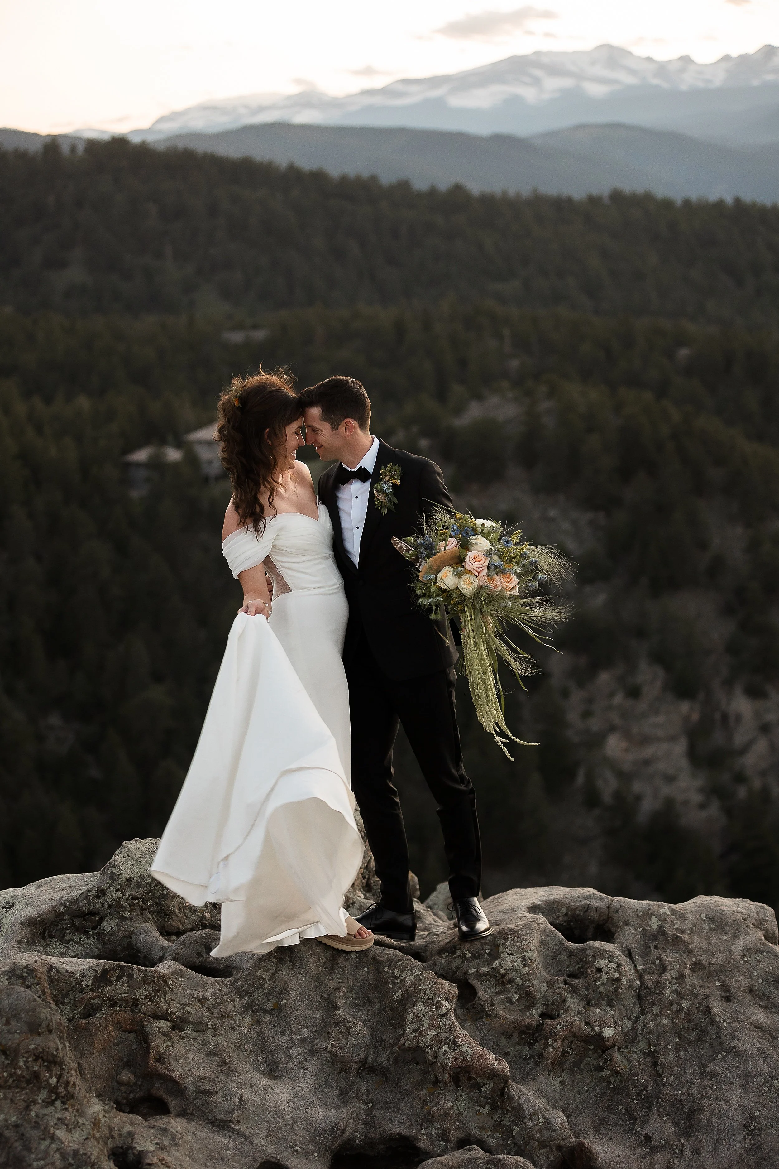 photo of a smiling bride and groom with their foreheads together on a rocky mountainside. the bride holds her dress in one hand the the groom holds her bouquet.  the background has distant snow-capped peaks at sunset