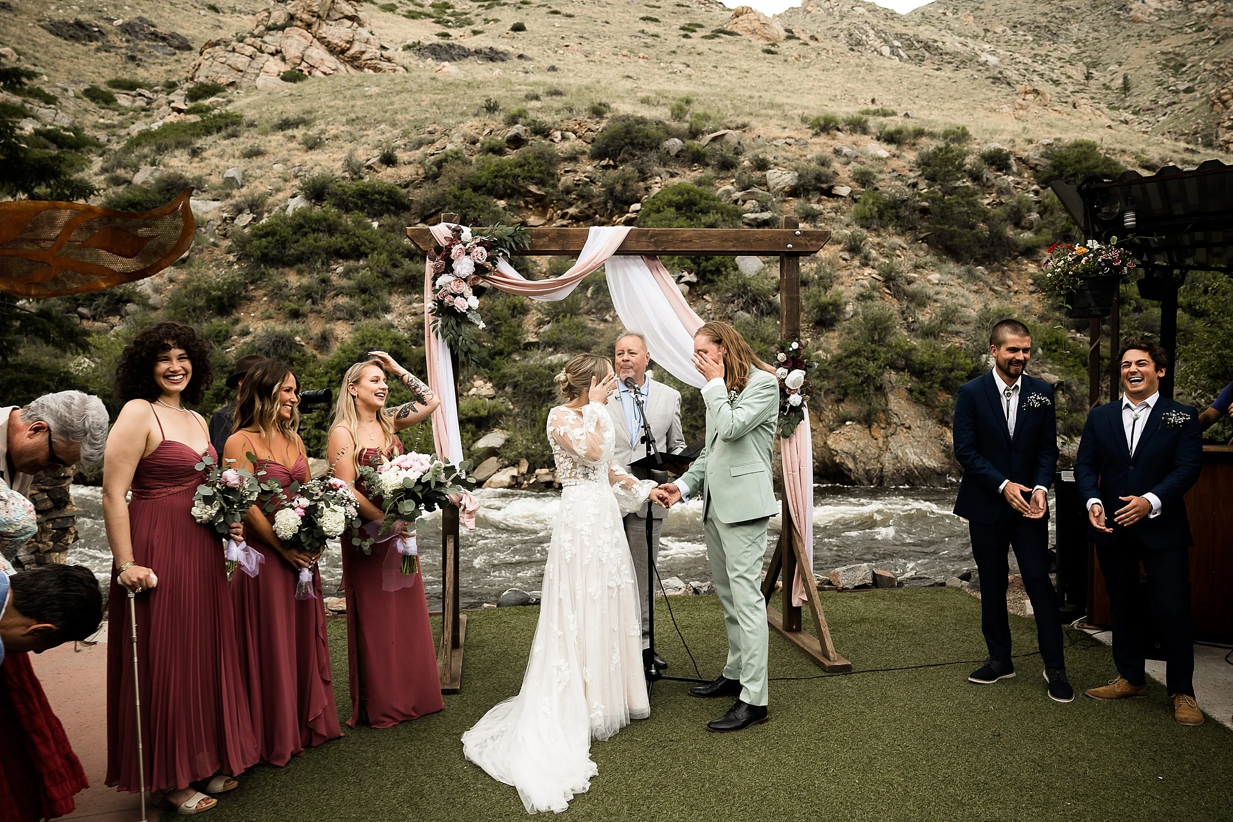 wedding couple wipes tears from their eyes during an emotional ceremony with a river and mountain backround