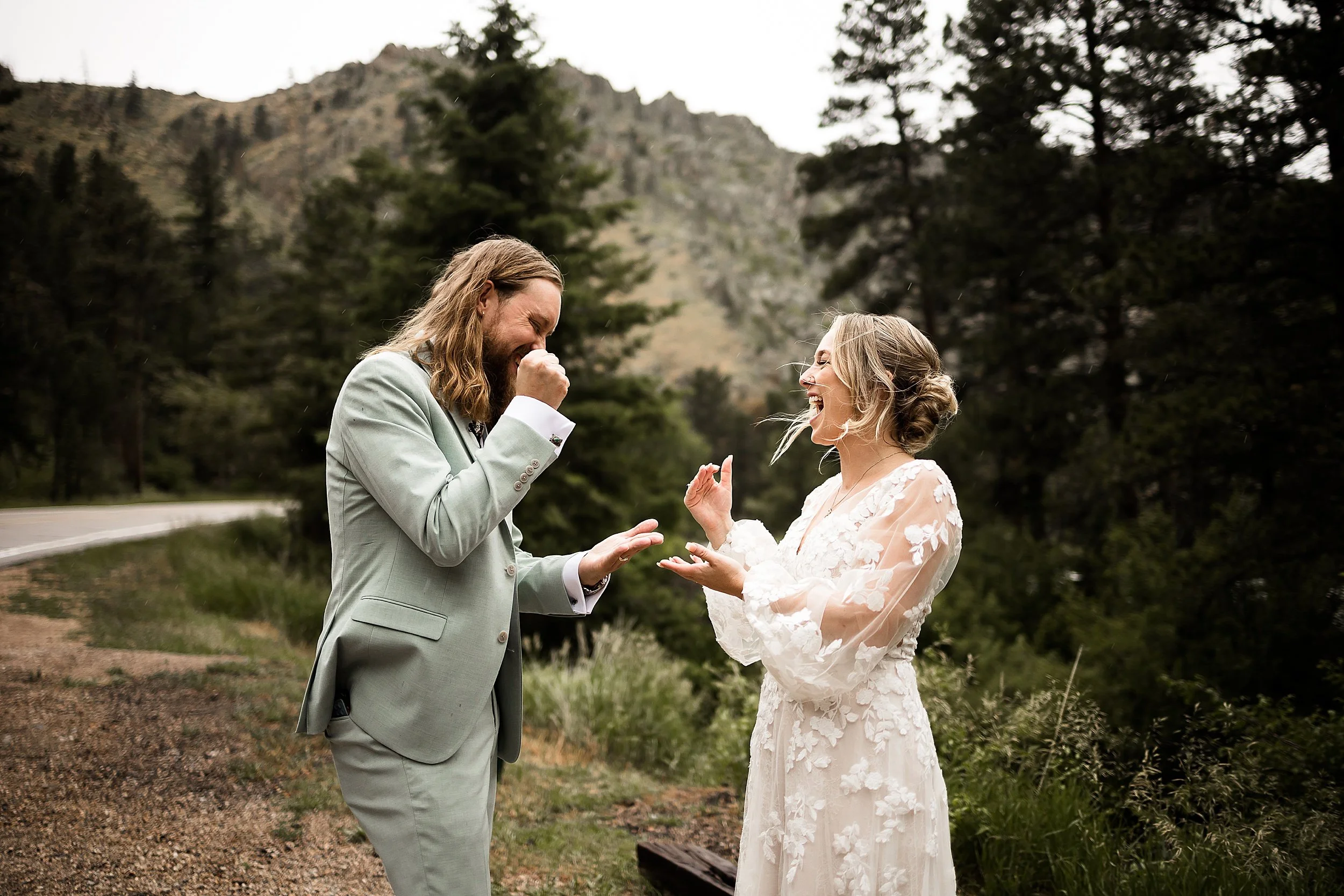 wedding couple plays rock paper scissors, both are smiling, there is a backdrop of pine trees and mountains