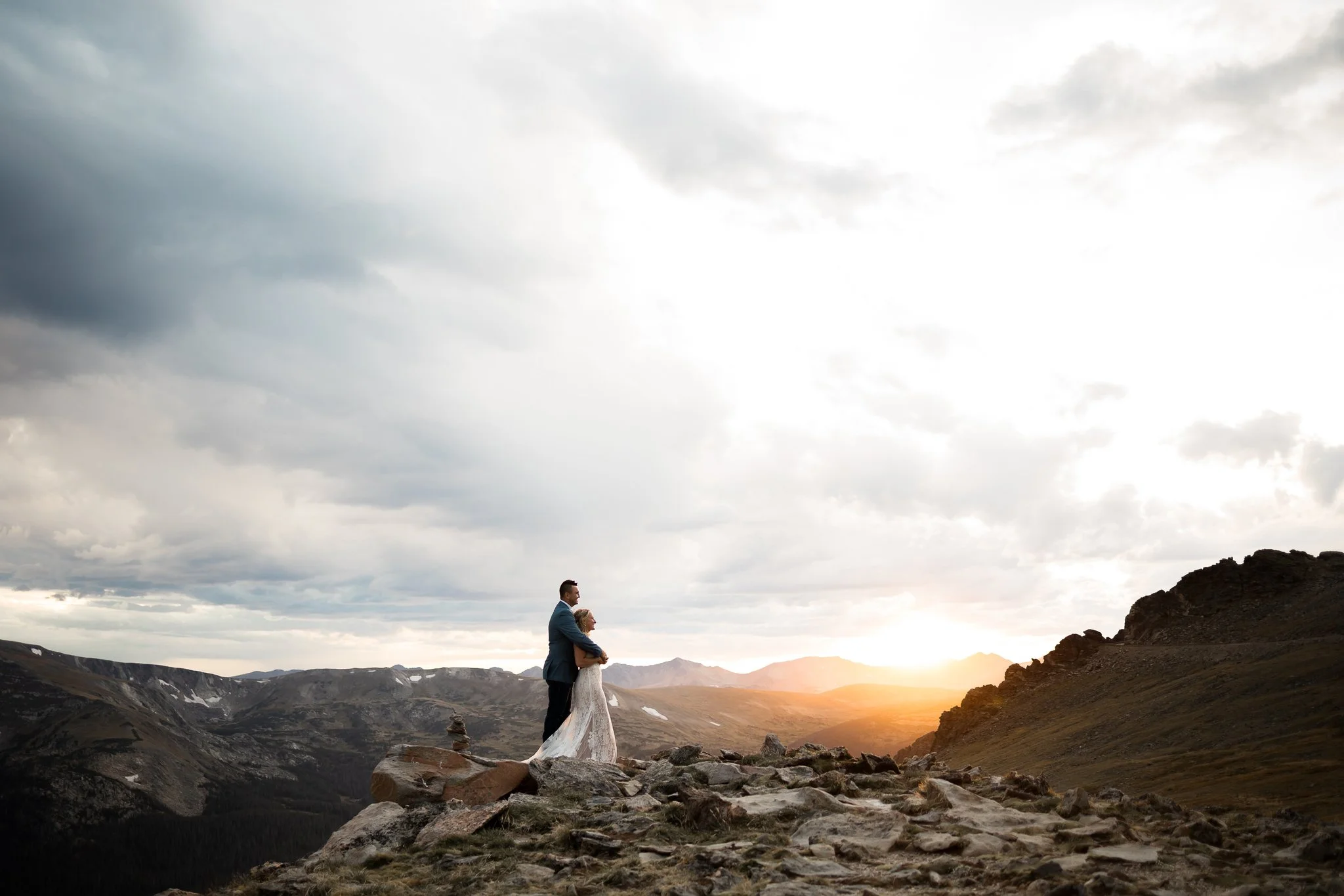A couple in wedding attire standing on rocky terrain in a mountainous landscape at sunset.