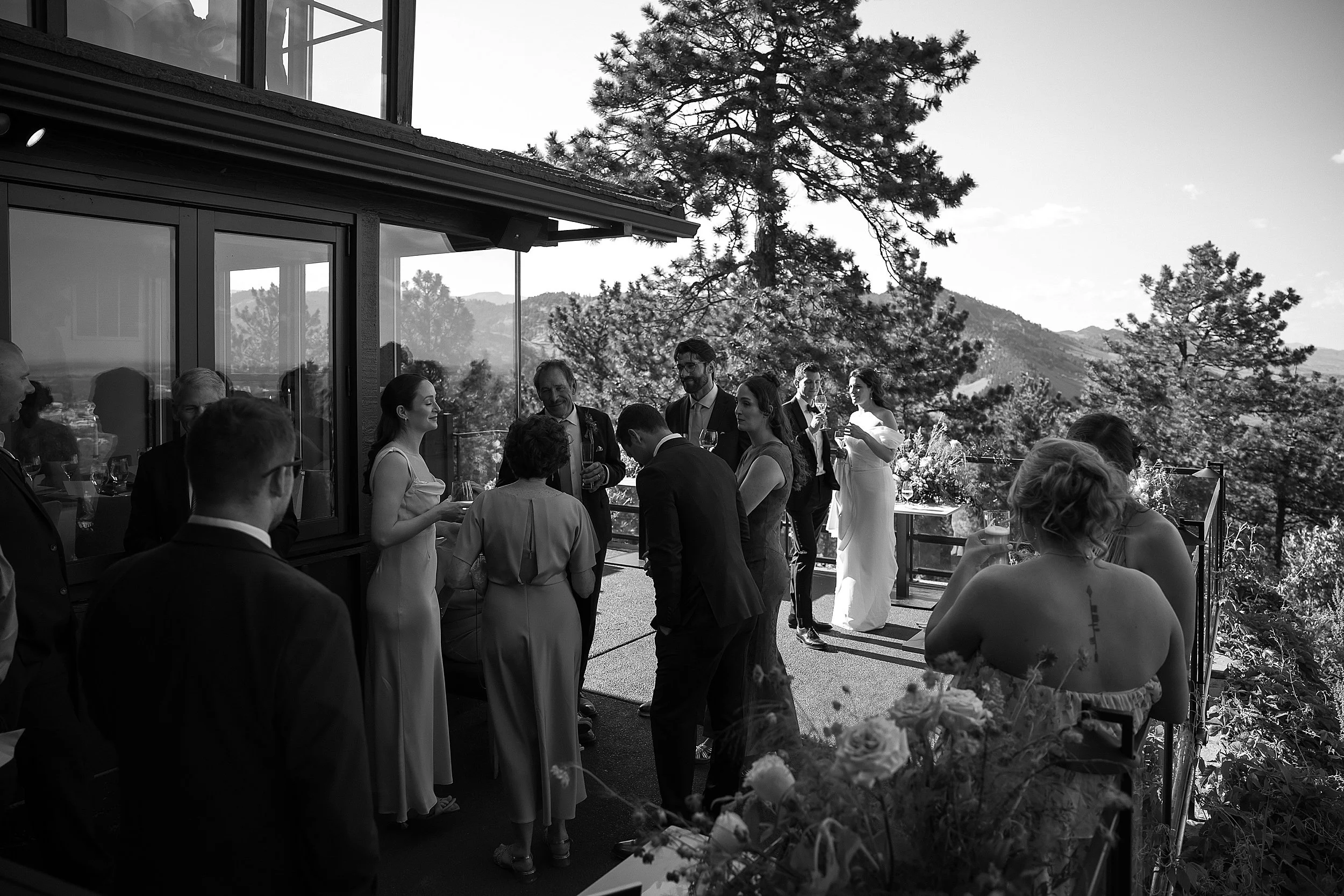 black and white image of a small group gathers on a terrace. they can be seen talking and drinking wine. a bride and groom are in the background.