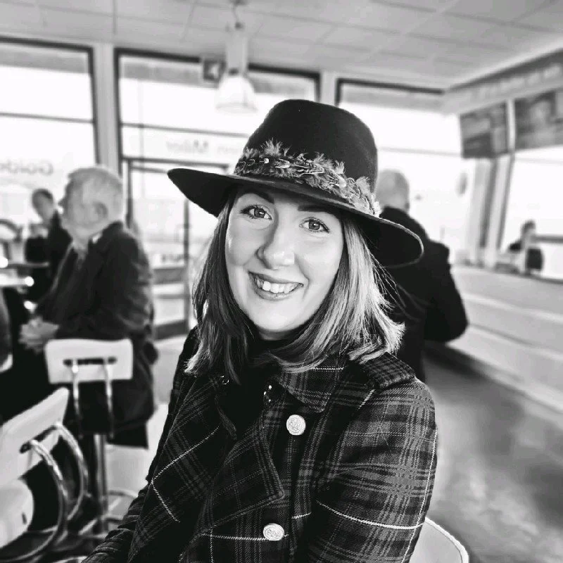 A smiling woman with shoulder-length hair wearing a wide-brimmed hat and plaid jacket, sitting in a cafe or similar setting.