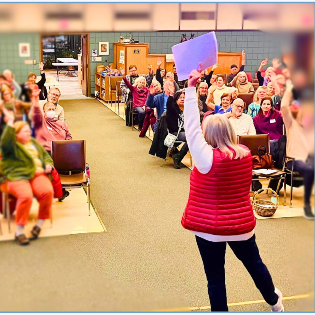 Members raising their hands along with the speaker at a meeting.