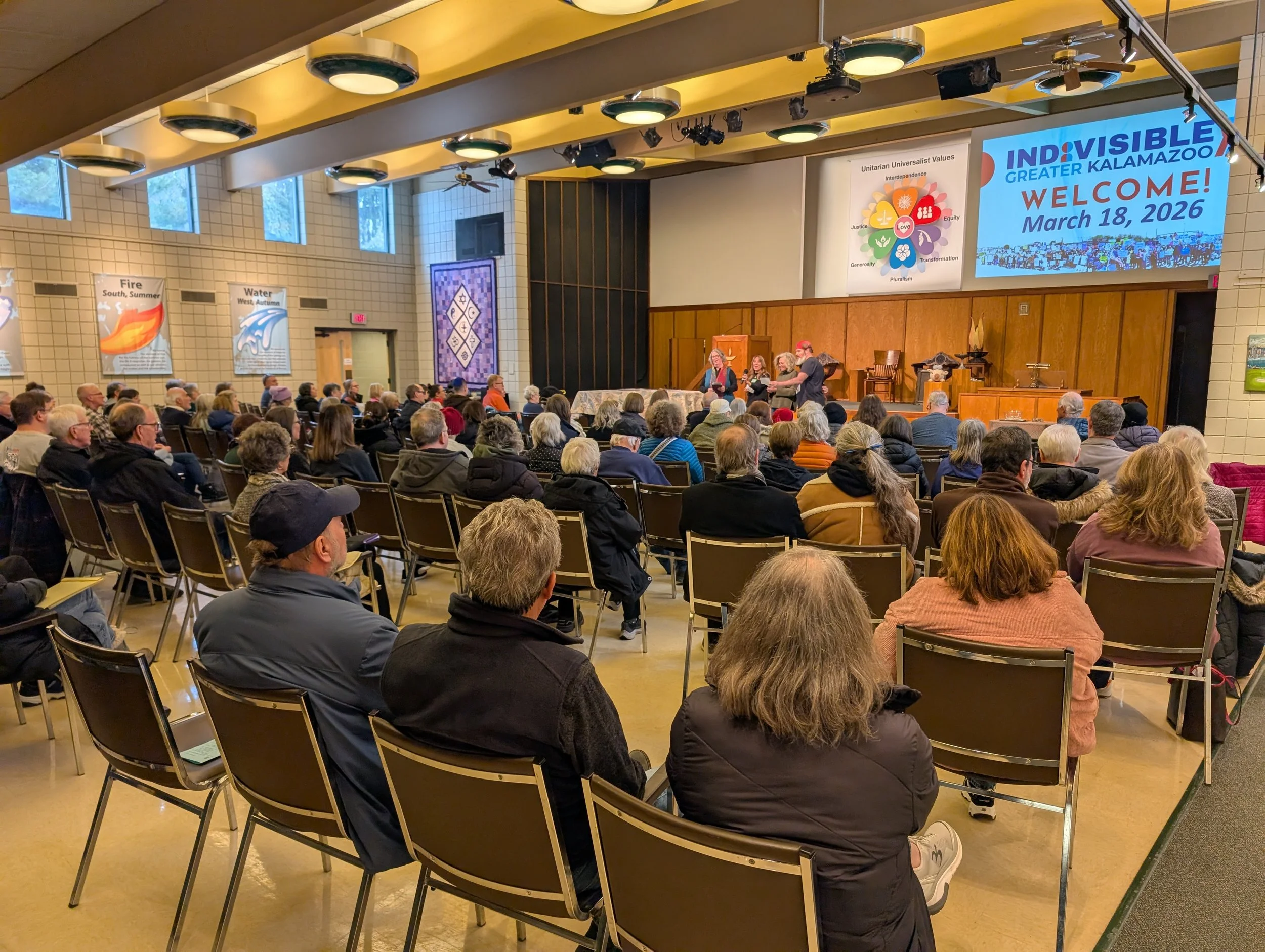 A large group of people seated in a conference room listening to a presentation. The stage at the front has a panel of speakers and a large screen displaying the text "INVISIBLE GREATER KALAMAZOO WELCOME! March 18, 2026". The room has high windows and posters on the walls.