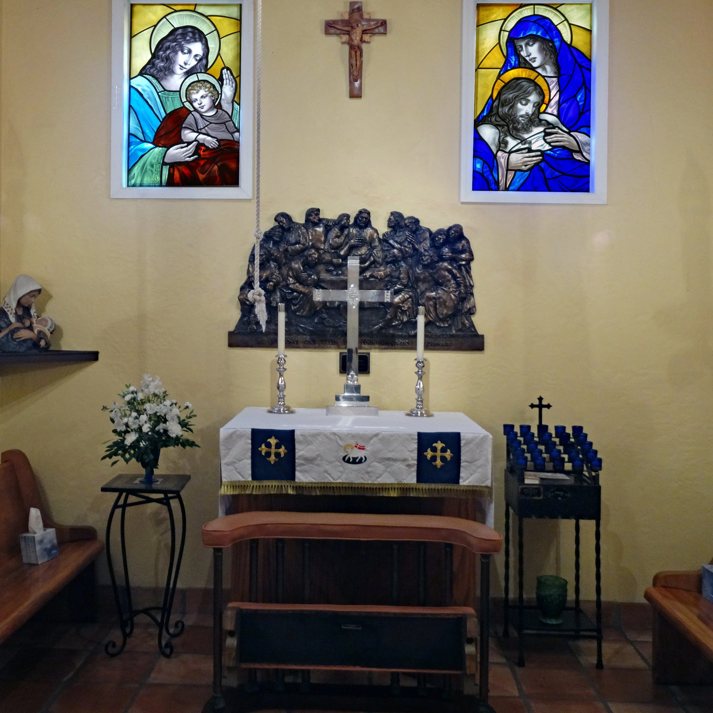 An altar in a church with stained glass windows, a crucifix, and religious statues. The altar has candles, flowers, and religious symbols.