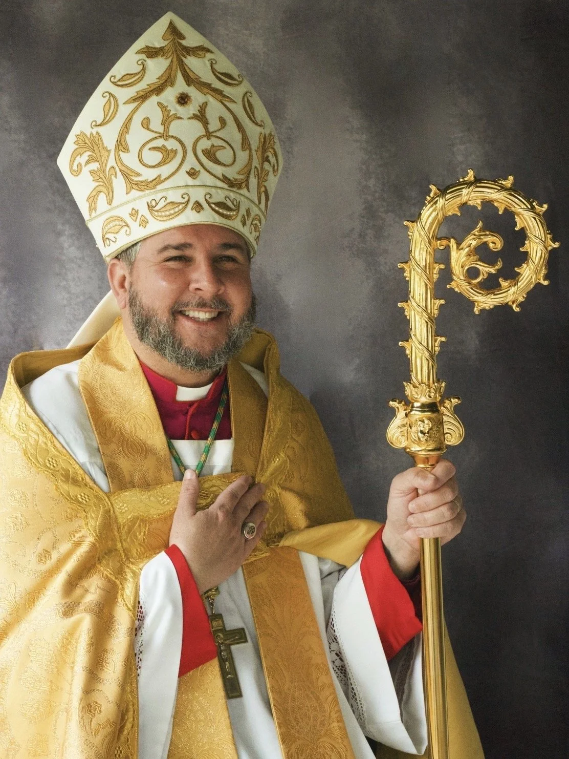 Man dressed as a bishop or pope, wearing a gold and white mitre, gold vestments, holding a gold staff, standing against a dark background.