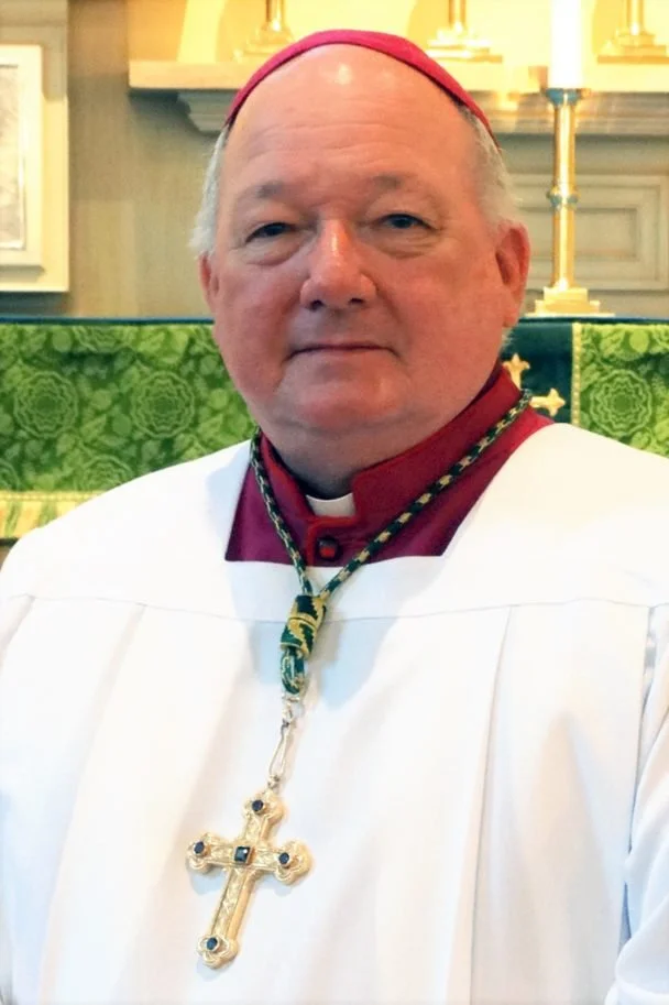 A man dressed in religious attire, wearing a red clergy hat and a large ornate cross necklace, standing in front of a church altar with lit candles.