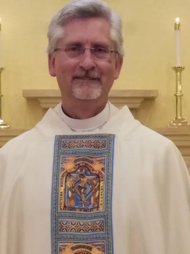 A priest with glasses and gray hair standing in front of a yellow wall with candles, wearing a white stole with a detailed religious design.