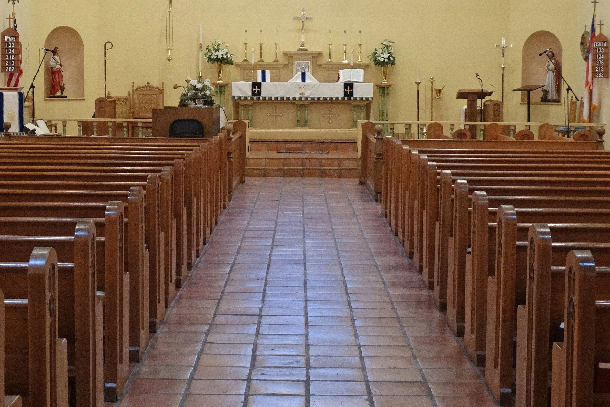 The interior of a church with wooden pews, an altar decorated with flowers, candles, and religious symbols, and statues on either side.