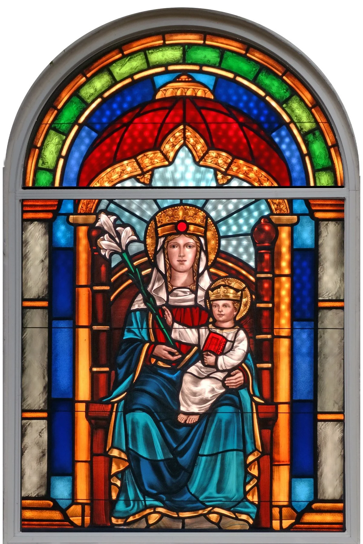 Colorful stained glass window depicting the Virgin Mary holding a lily and a child, both with halos, seated on a throne with a red book in her lap. The scene is framed by an arched window with vibrant colors and ornate details.