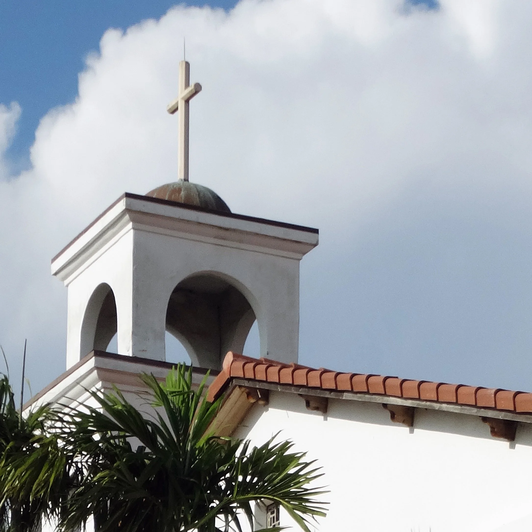 A church steeple with a white cross on top, against a partly cloudy sky.