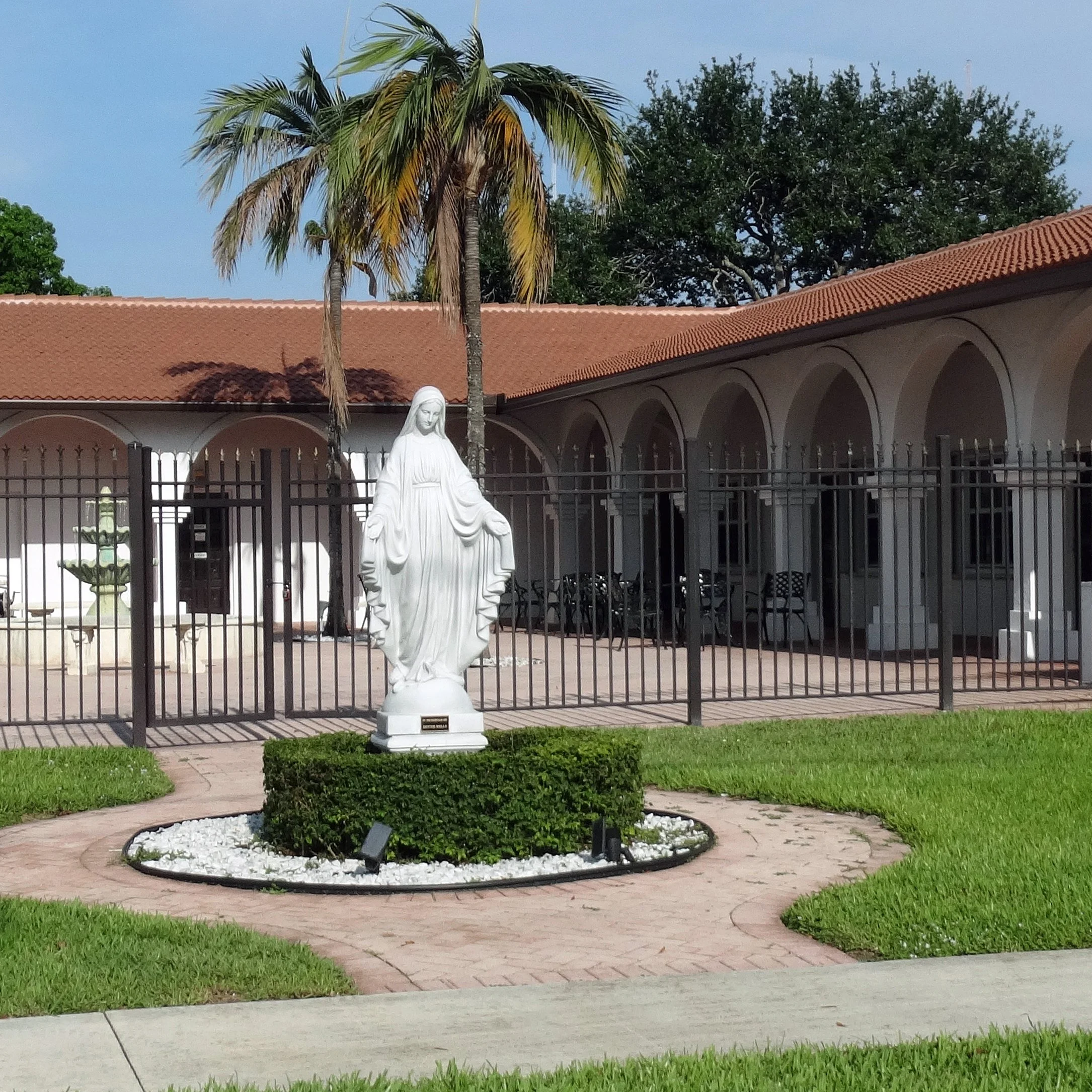 A white statue of the Virgin Mary standing on a small, circular, landscaped area with white rocks and green bushes, surrounded by a paved walkway. Behind the statue is a black fence and a building with arched walkways and a red-tiled roof, with a palm tree and other trees visible in the background.