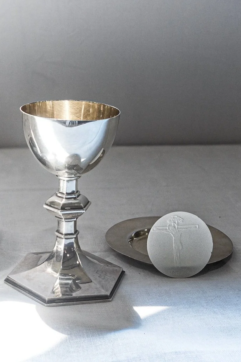 A silver chalice and a white communion wafer on a silver dish, set on a white tablecloth with a gray background.