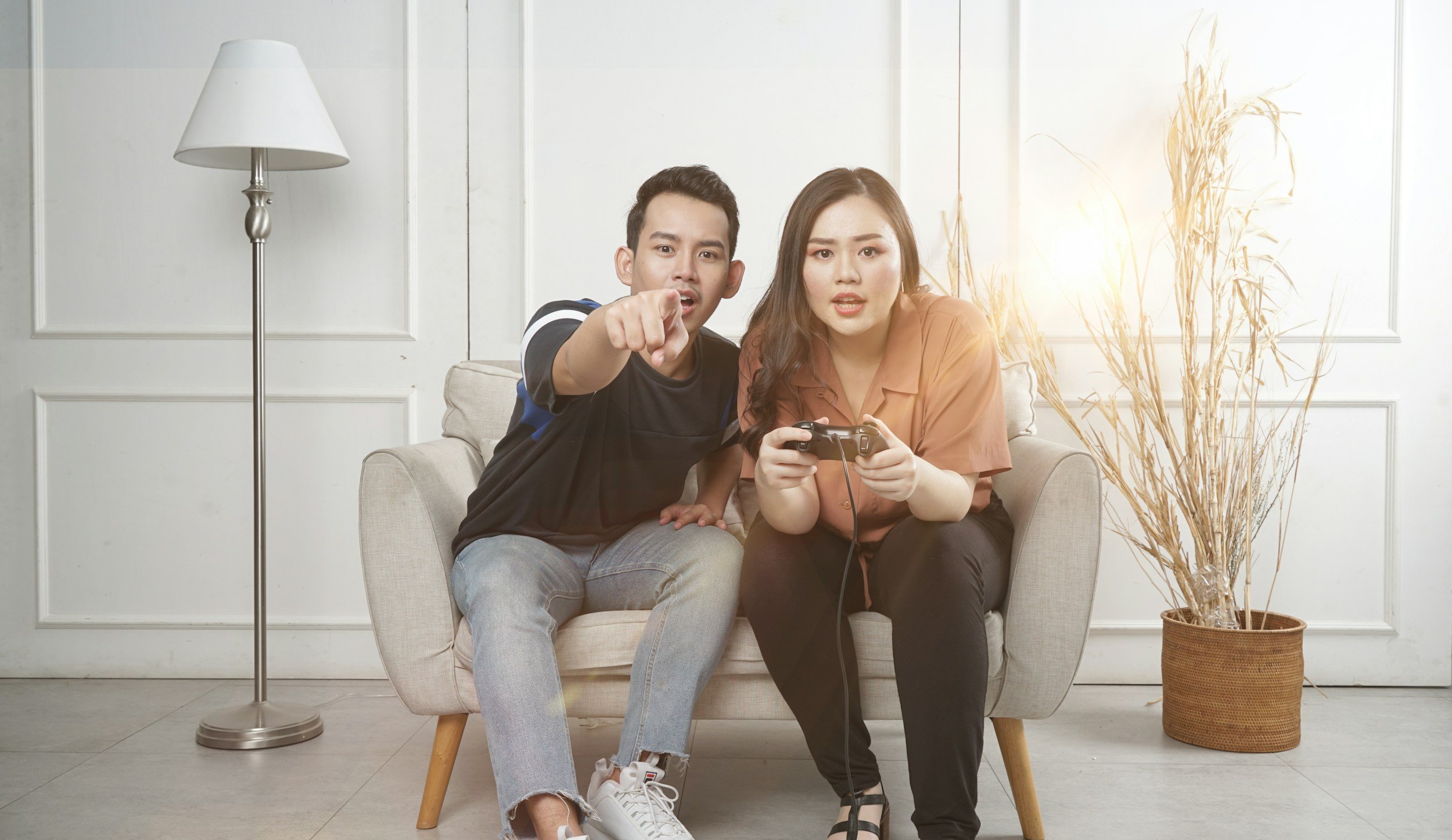 Young man and woman sitting on a beige sofa in a bright room, with the man pointing at the camera and the woman playing a video game.
