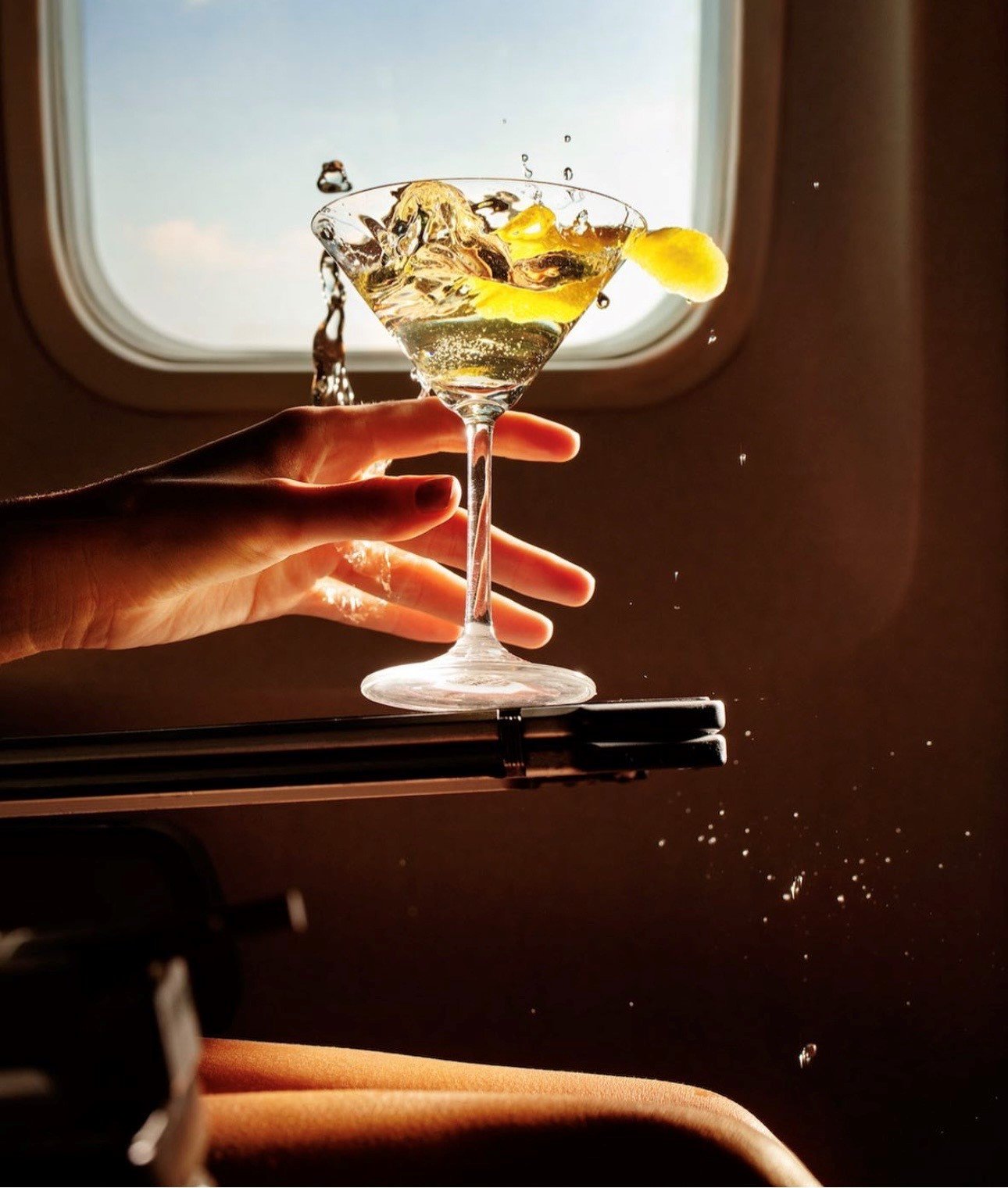A hand holding a martini glass filled with a yellowish drink, garnished with a lemon slice, with water splashing out of the glass, set against an airplane window.