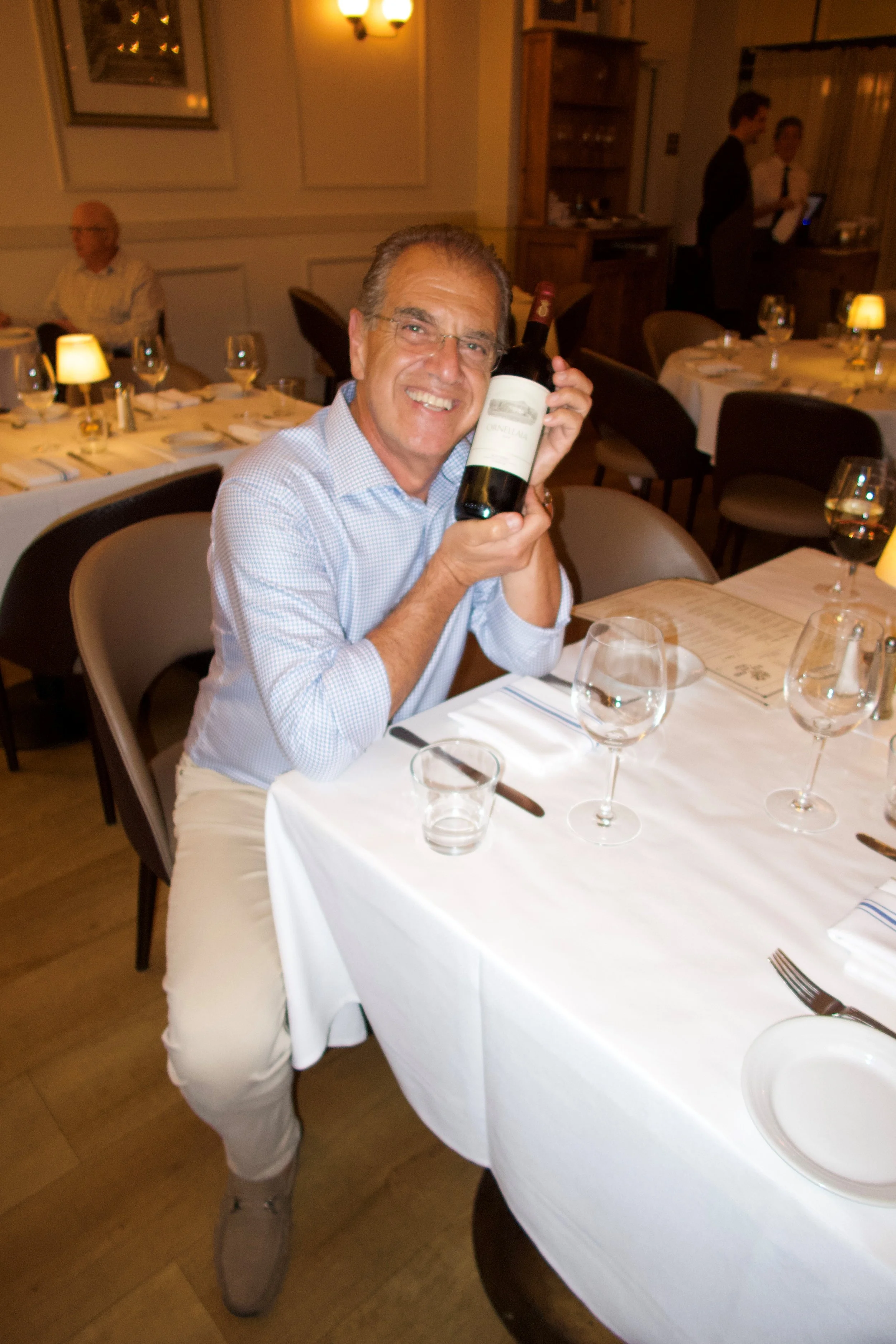 A man with glasses, wearing a checkered shirt and light-colored pants, is smiling and holding a bottle of red wine in a restaurant setting with tables, wine glasses, and dim lighting.