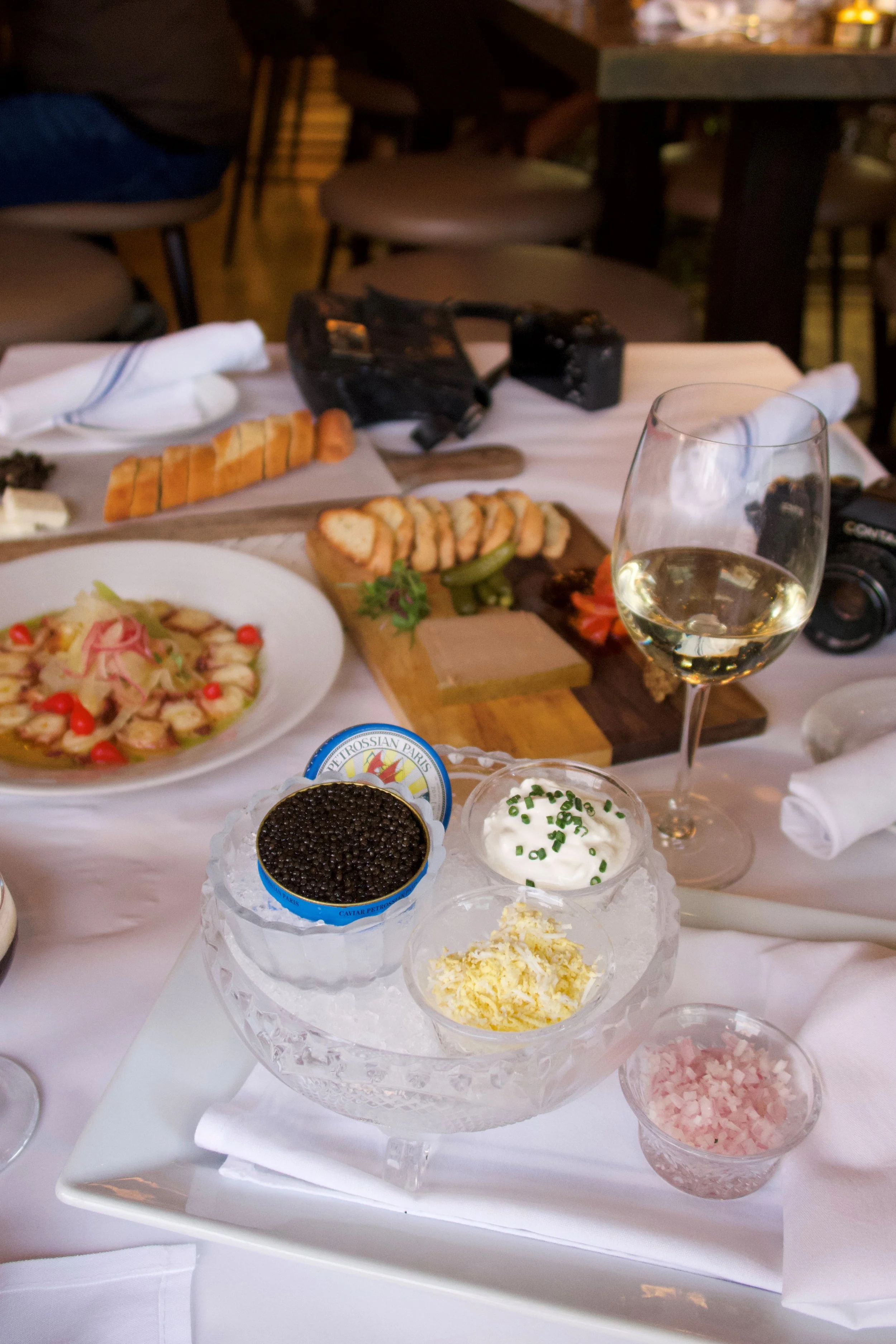 A table set with various dishes and condiments, including a bowl of caviar, chopped onions, and creamy dips, with a glass of white wine in the foreground and a camera on the table. The background shows a dining area with chairs and tables.
