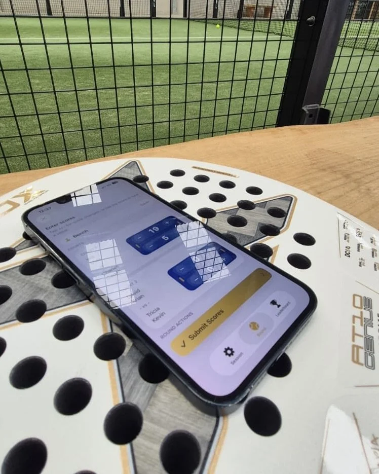 A smartphone displaying a score submission screen placed on a padel racket with court in the background