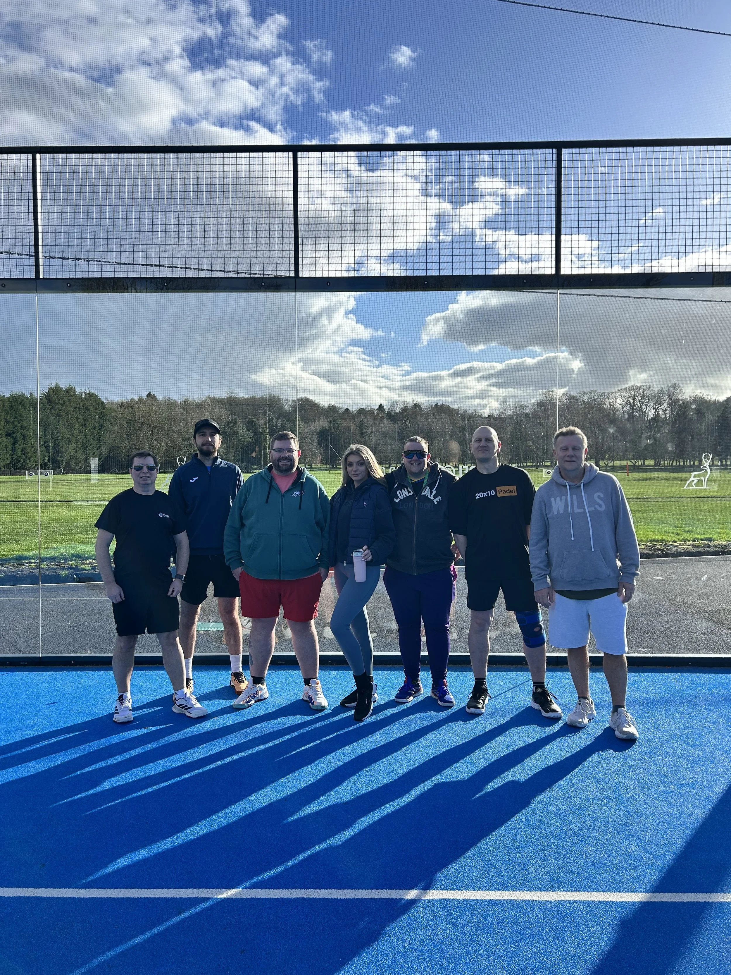 Group of seven people standing on an outdoor blue padel court, with a glass wall behind them and green field in the background. The sky is partly cloudy.