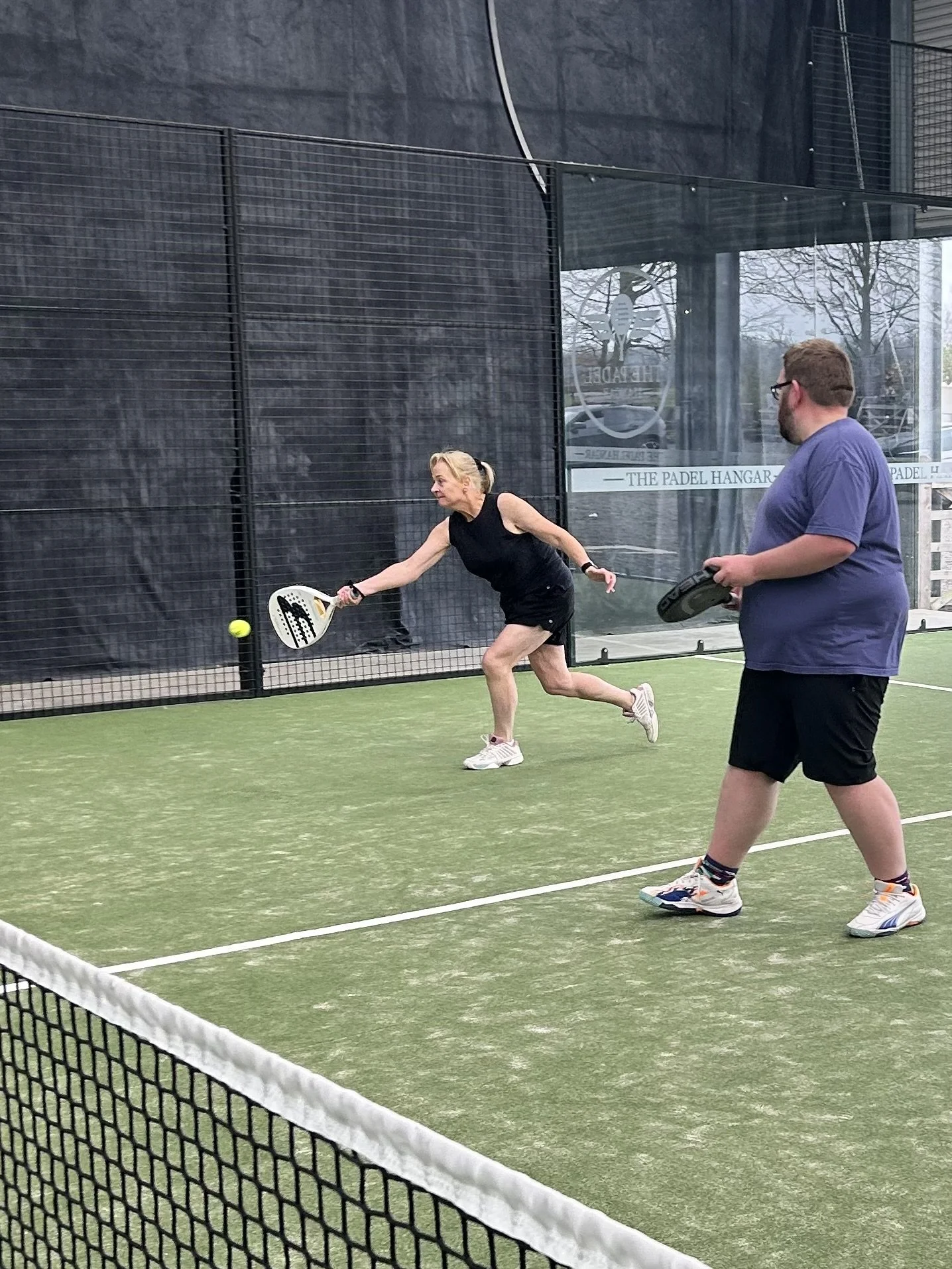 Two people playing padel on an indoor court. The woman is hitting the ball while the man watches, holding his racket.