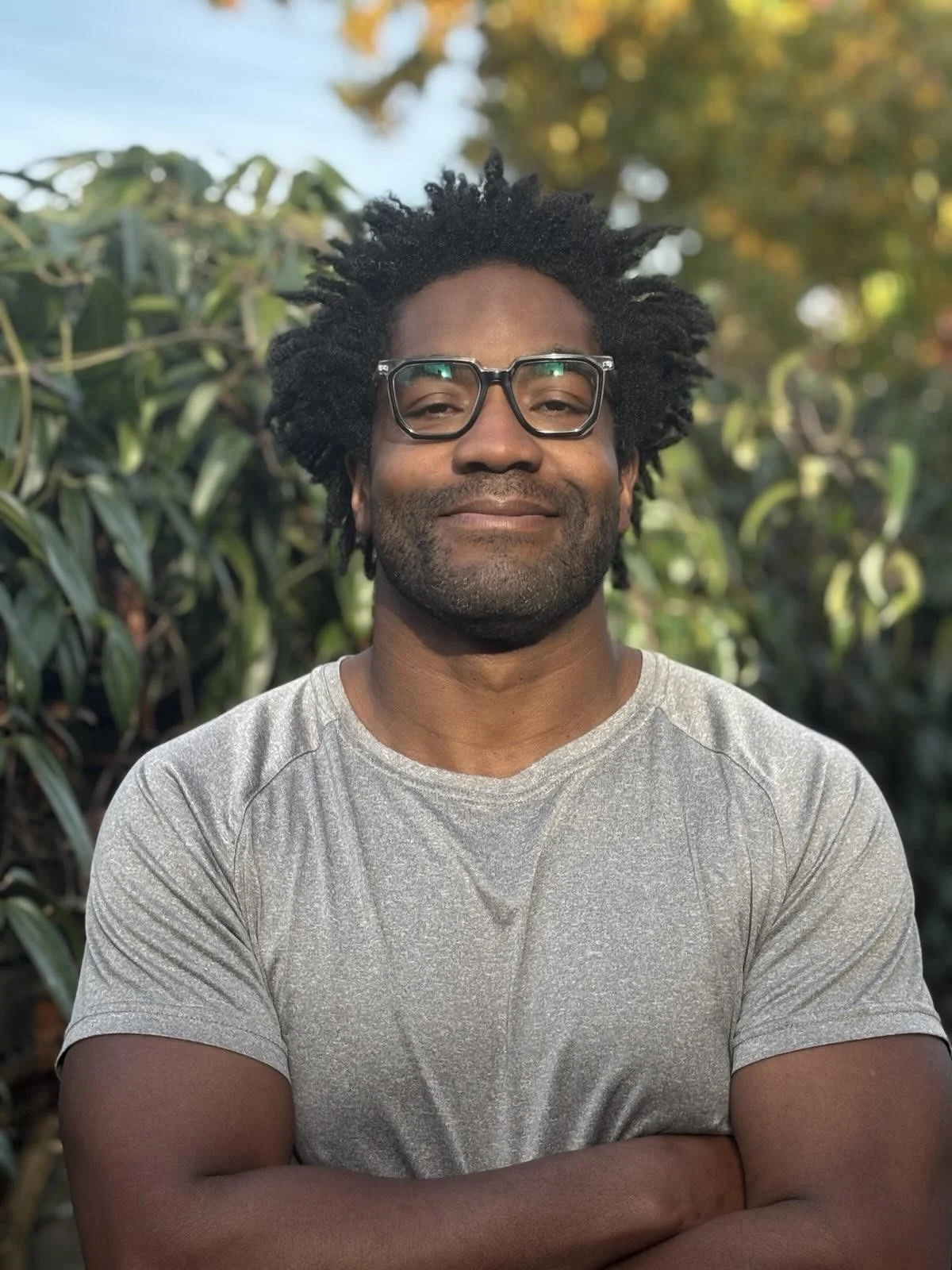 A man with glasses and a beard in a gray t-shirt standing outdoors in front of green plants and trees, smiling with arms crossed.