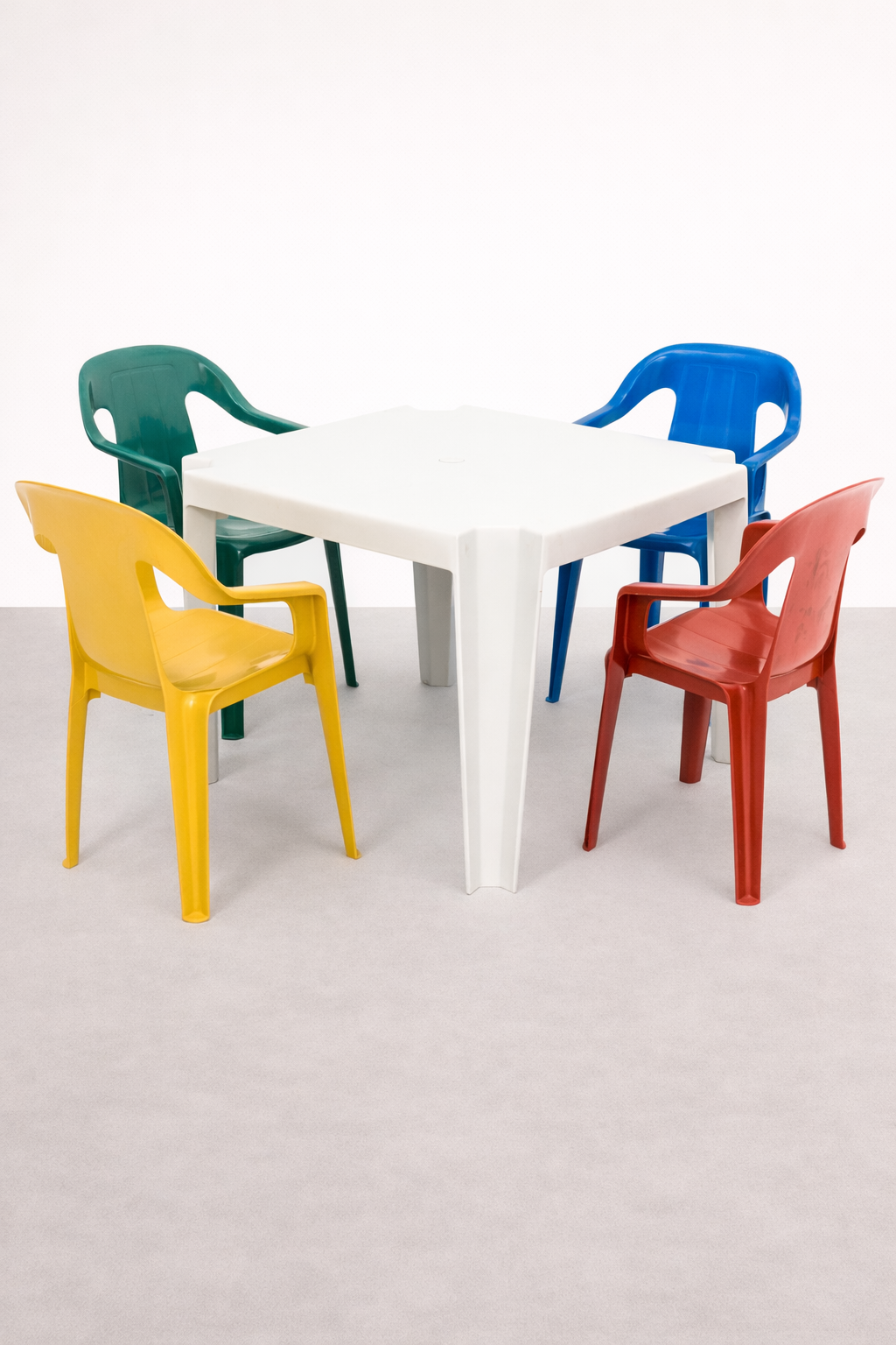Mesa branca com quatro cadeiras de plástico coloridas: uma amarela, uma verde, uma azul e uma vermelha, em um ambiente com parede branca e piso cinza.