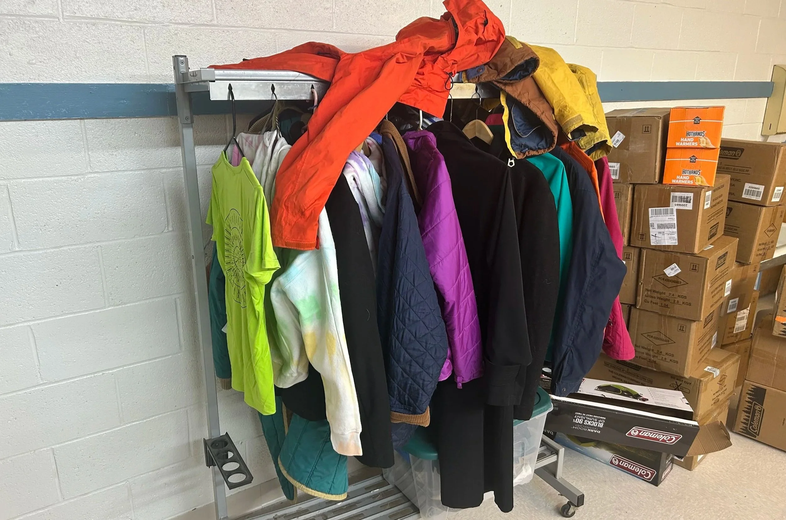 Clothing rack with jackets, hoodies, and shirts in various colors, atop a metal shelf, next to boxed items in a storage room.