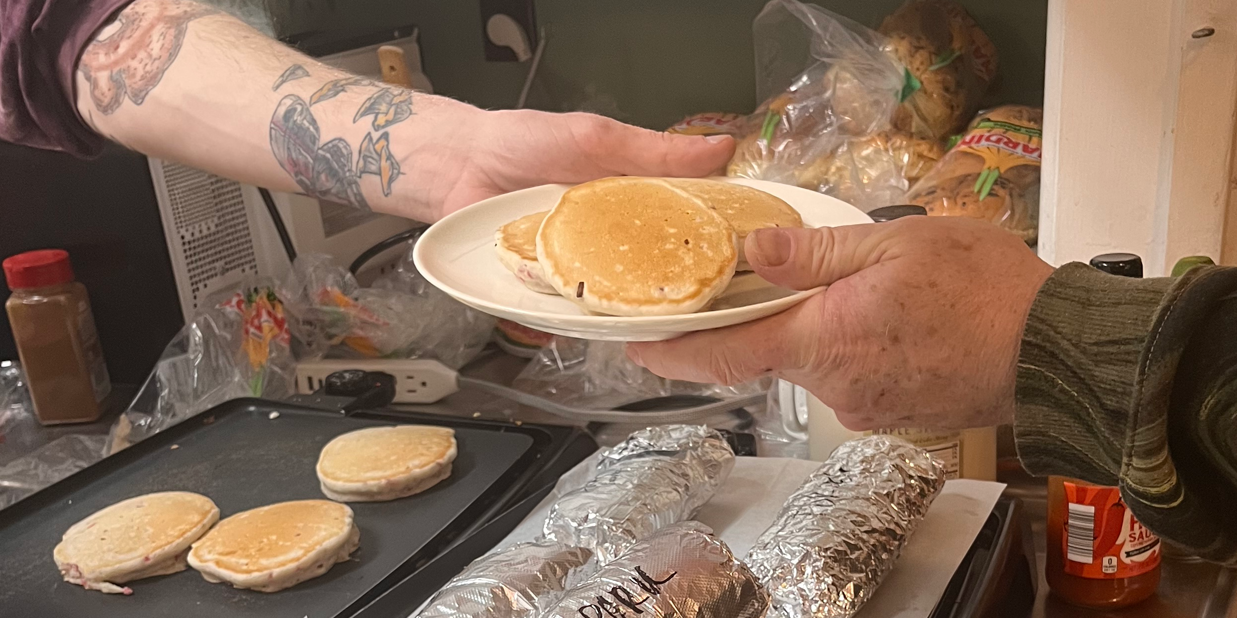 One person hands another a plate of pancakes. Only their hands and the plate are in the frame. There are four more pancakes on a black tray and several foil-wrapped burritos on a kitchen counter.