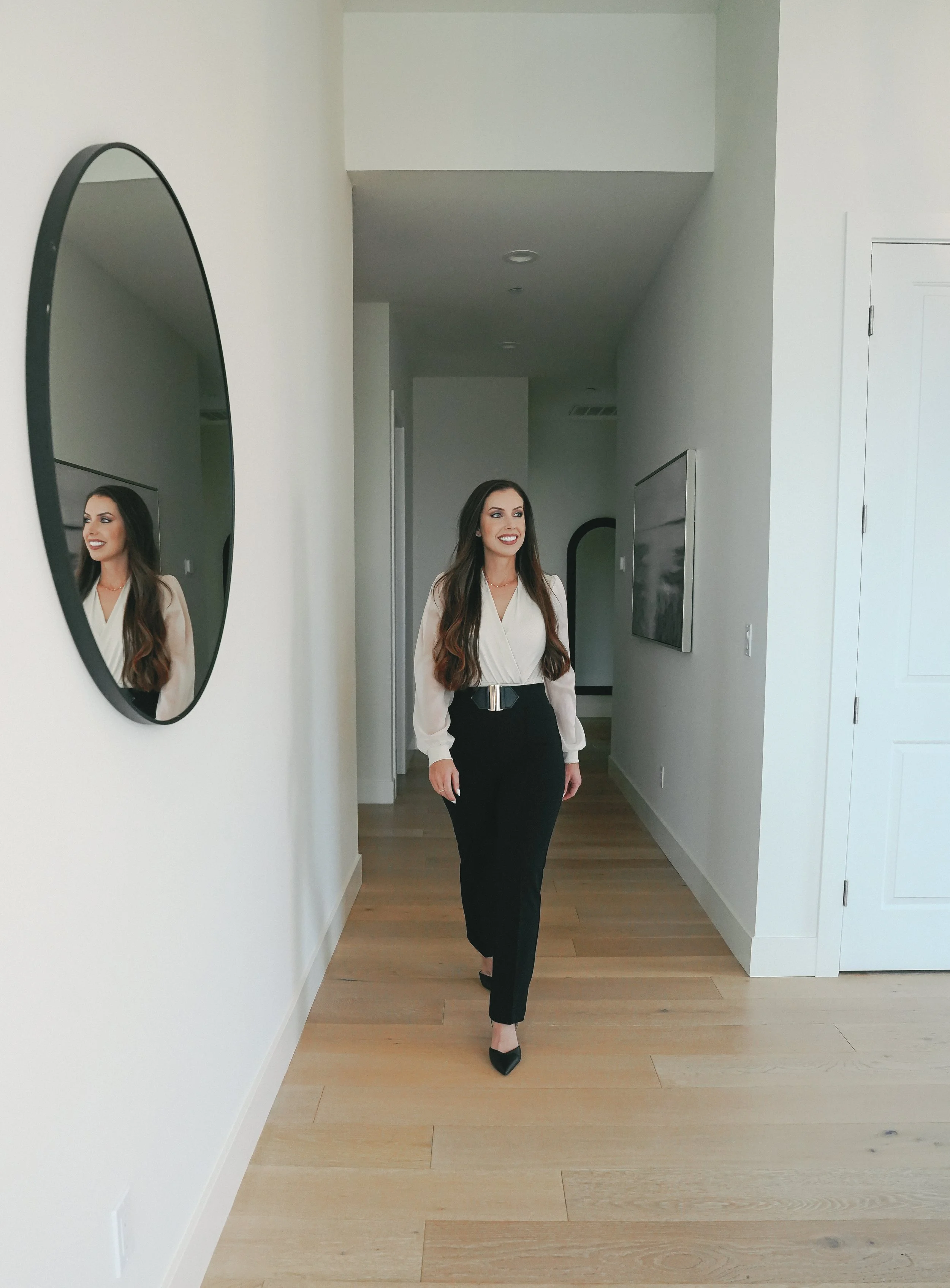 A woman with long hair wearing a cream blouse and black pants walking in a modern, minimalist home with light wood flooring, a large mirror on the wall, and a TV mounted on the wall.