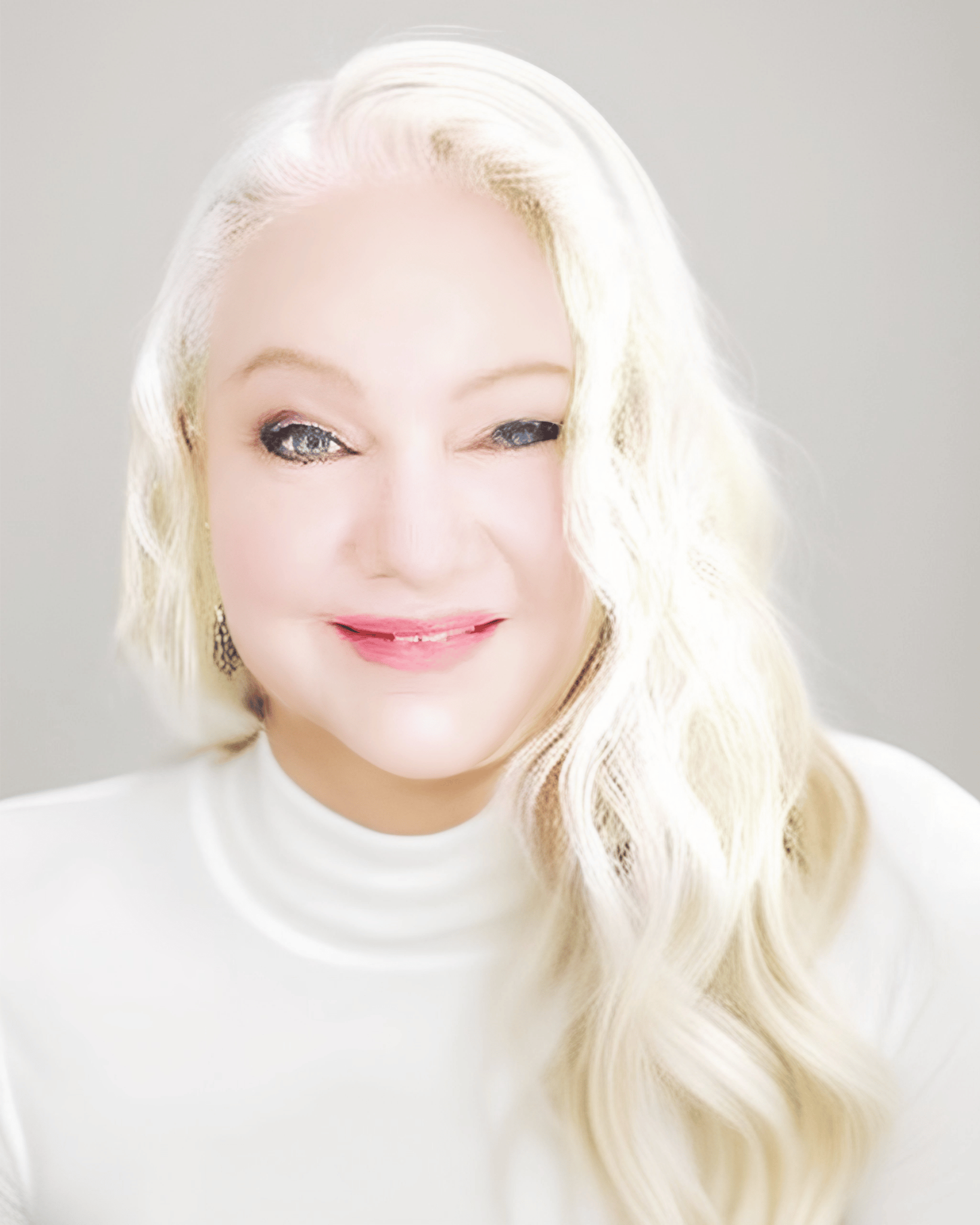 A woman with platinum blonde hair styled in loose waves, wearing a white top, smiling softly, with a light-colored background.