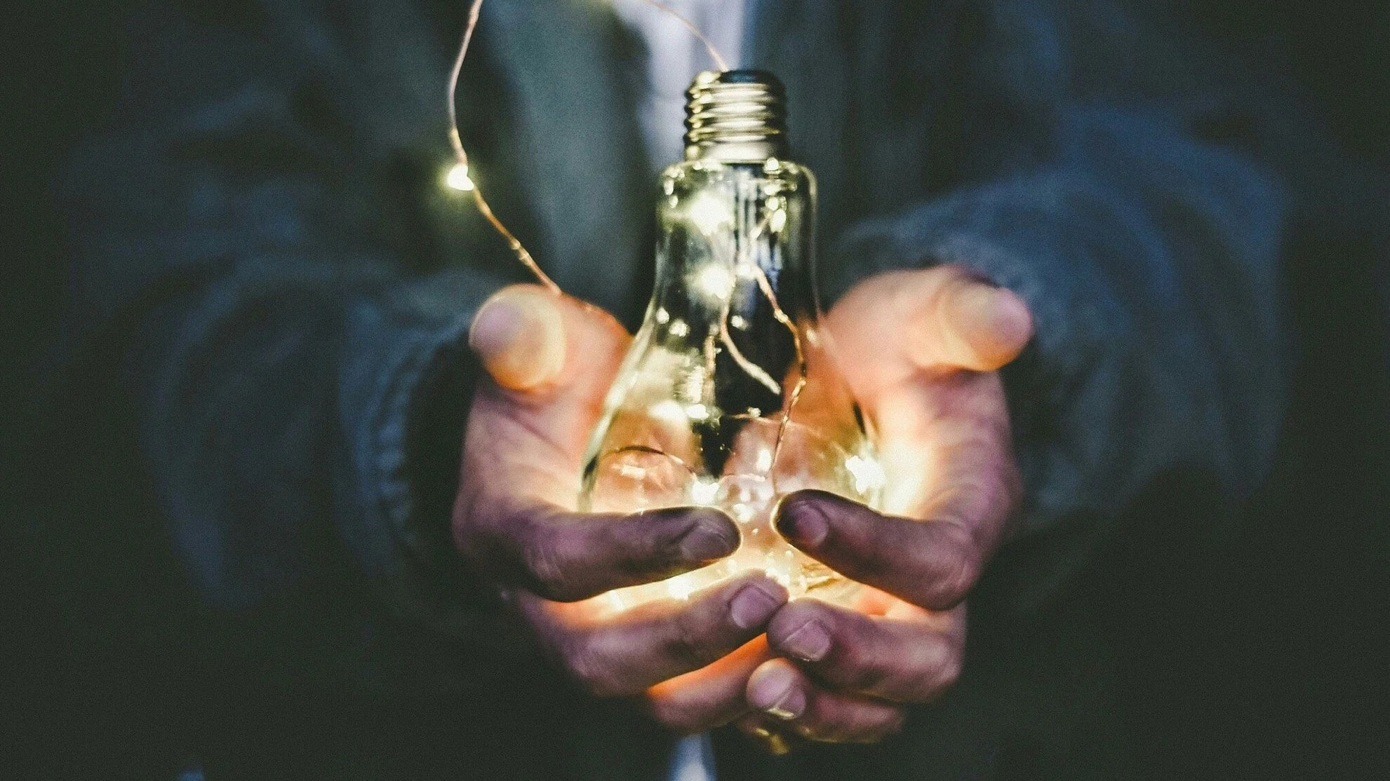 Two hands holding a lightbulb that contains string lights inside, with some of the lights wrapped around the person's fingers.