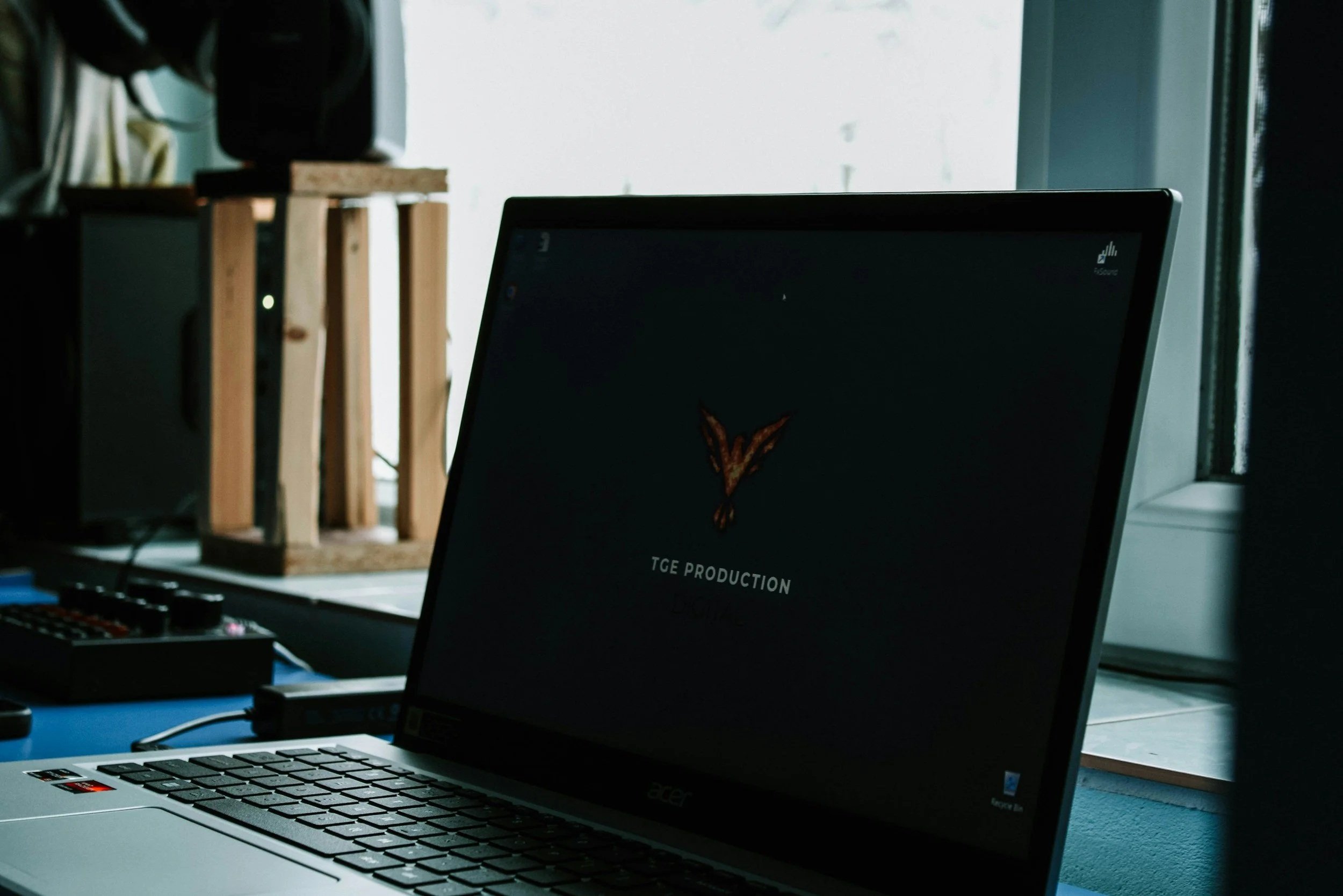 A laptop on a desk near a window, with a dark screen displaying a logo and the text 'TGE PRODUCTION.'