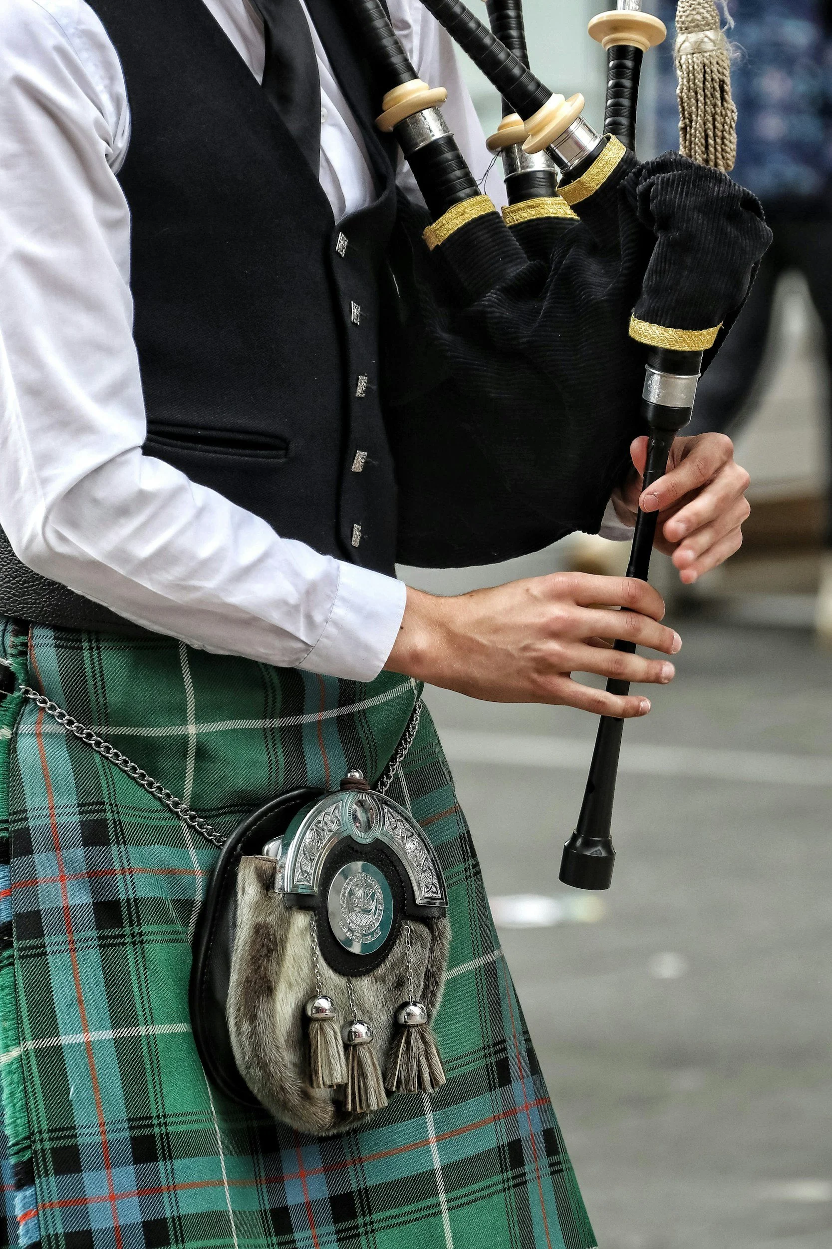 A person playing bagpipes, wearing a traditional Scottish kilt with a sporran, a vest, and a white shirt.