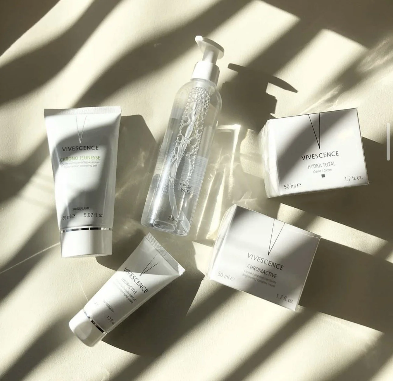 Produits de soin de la marque Vivesence, comprenant une crème, un gel nettoyant, une eau micellaire, et deux autres emballages, disposés sur une surface claire avec des ombres dessinées par la lumière.