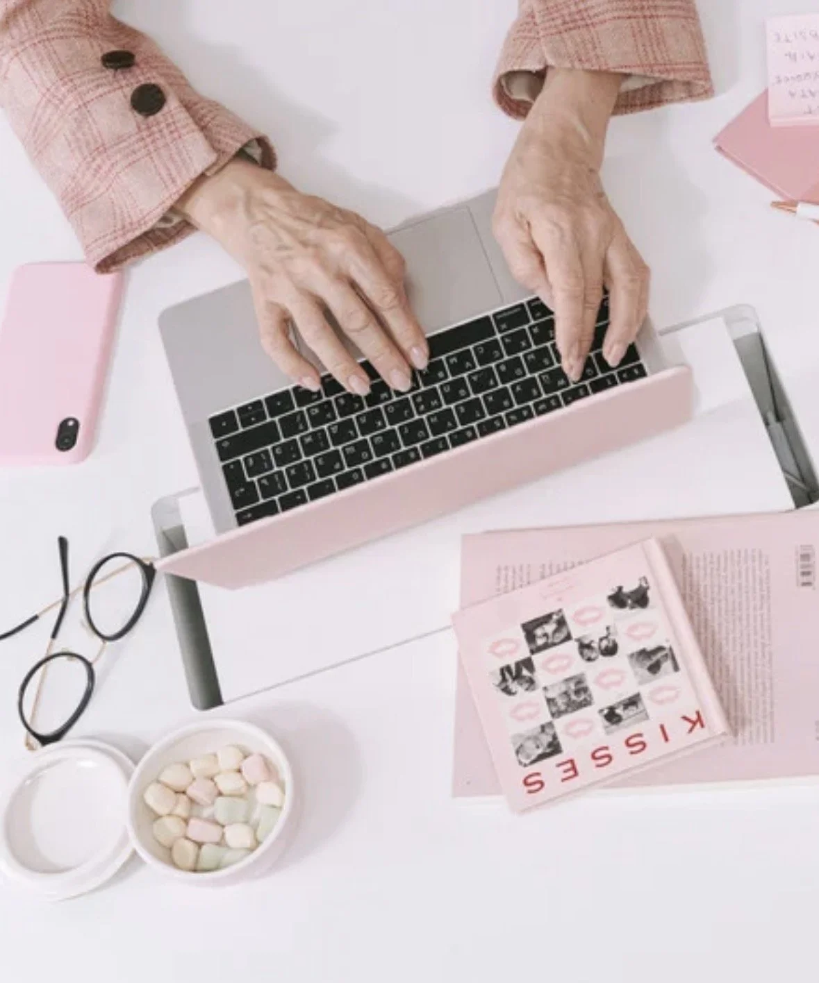 Une personne âgée tape sur un clavier d'ordinateur portable rose clair. Sur la table, il y a des lunettes, un téléphone rose, une petite boîte avec des bonbons colorés, et des brochures ou magazines roses.