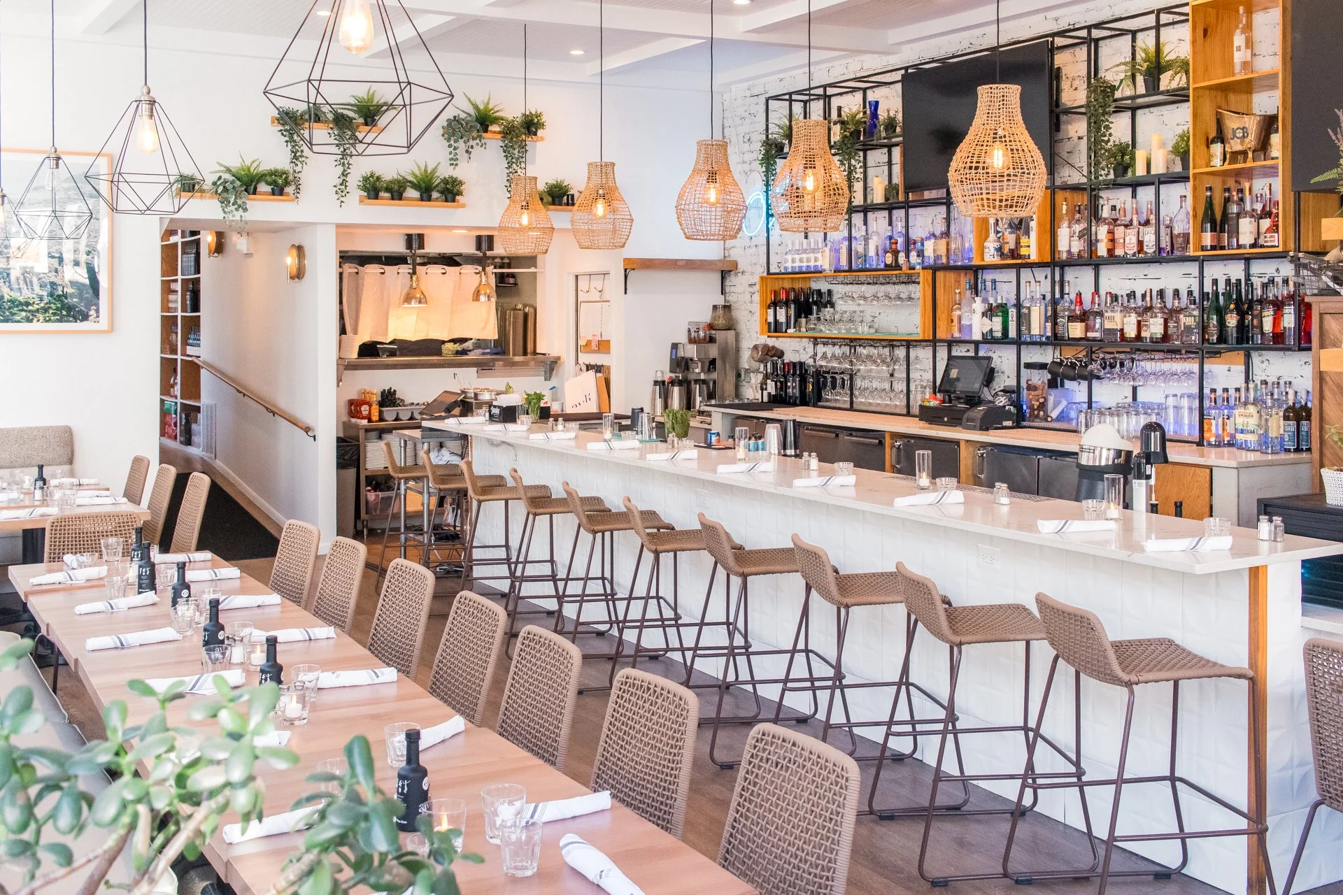 Modern restaurant interior with a bar, barstools, dining tables, woven pendant lights, and decorative plants.